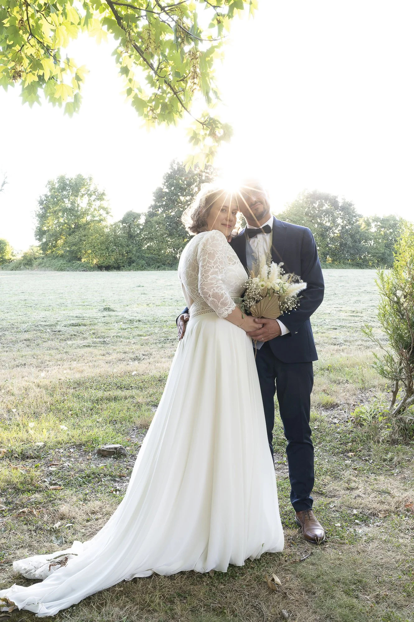 Un couple en costume de mariage debout dans un espace extérieur verts avec des arbres, tenant un bouquet de fleurs. La lumière du soleil brille derrière eux, créant un effet de halo.