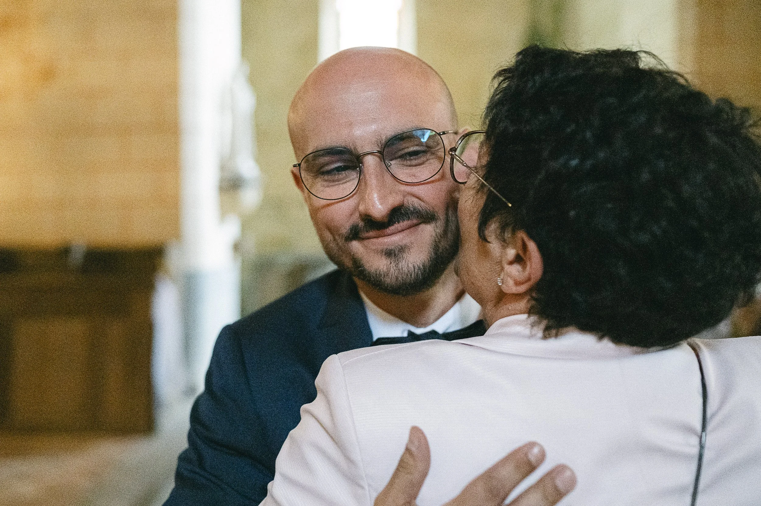 Un homme avec des lunettes sourit pendant qu'il serre dans ses bras une femme qui porte une robe beige, dans une scène chaleureuse et intime.