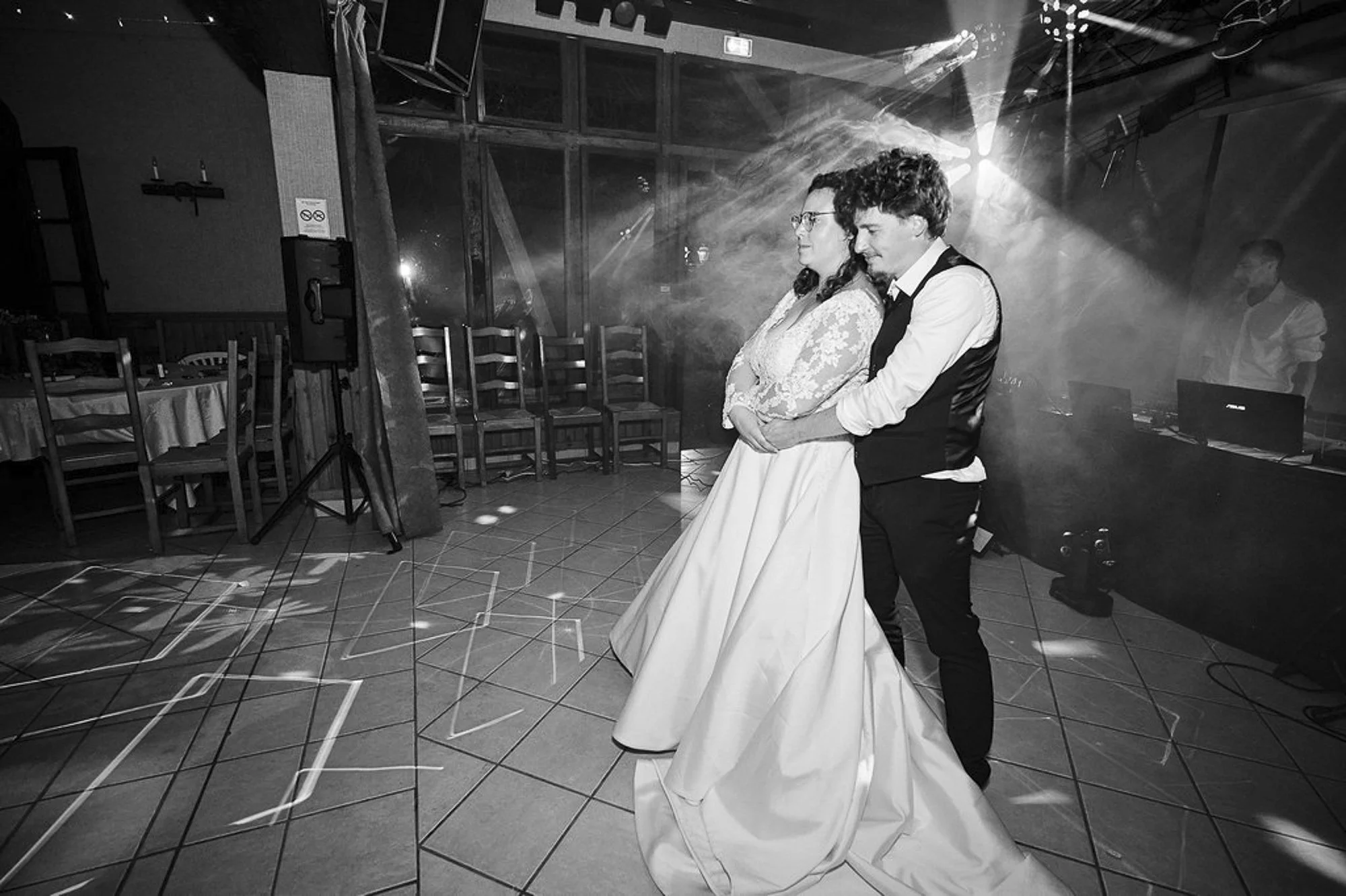 Un couple danse lors d'une célébration ou d'un mariage, en noir et blanc, dans une salle décorée avec des chaises en bois et des éclairages de fête.