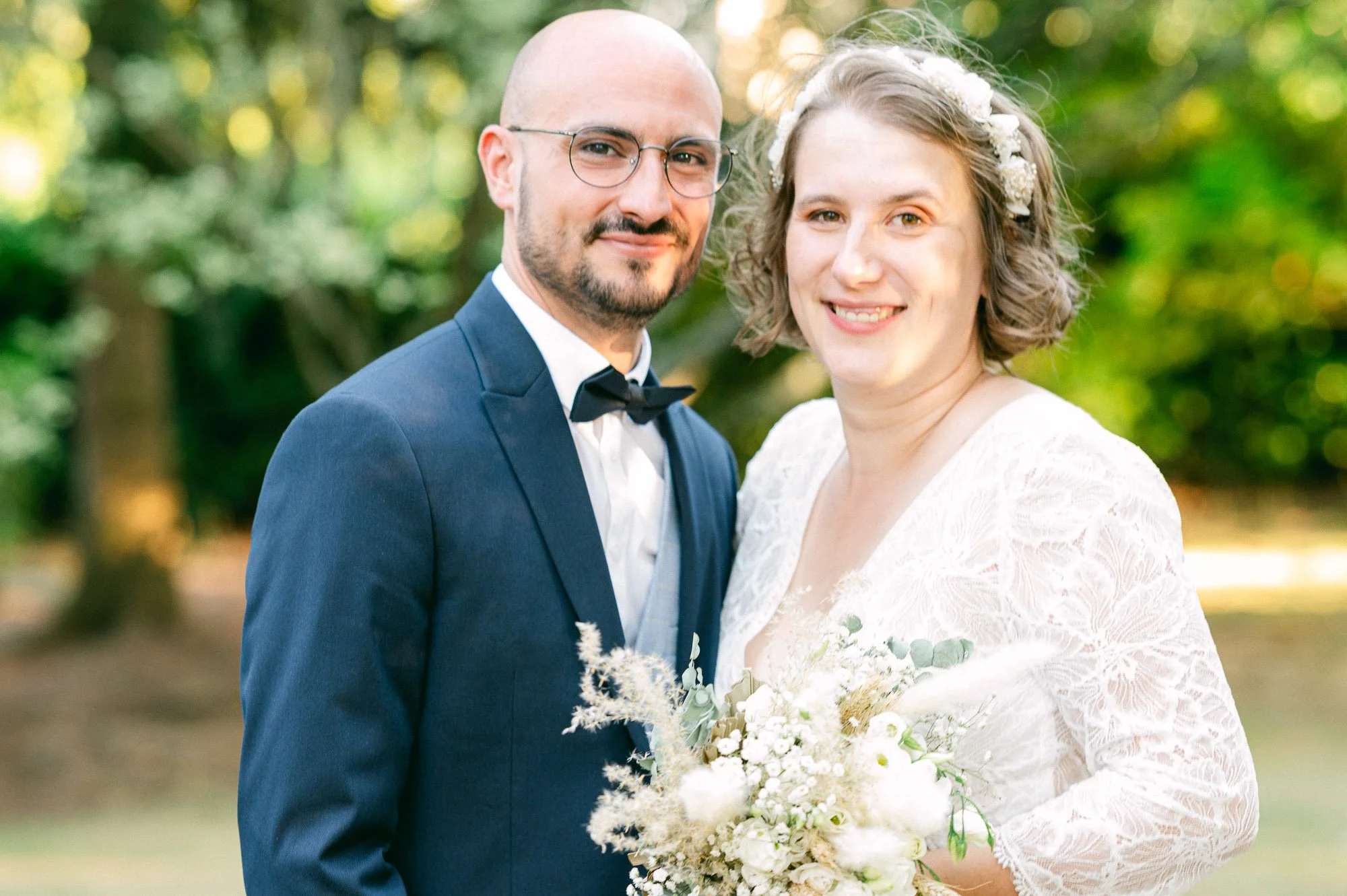 Un couple en mariage, l'homme porte un costume bleu avec une chemise blanche et une boucle d'oreille, la femme porte une robe blanche en dentelle avec une couronne de fleurs dans les cheveux, tenant un bouquet de fleurs blanches, dans un parc avec de