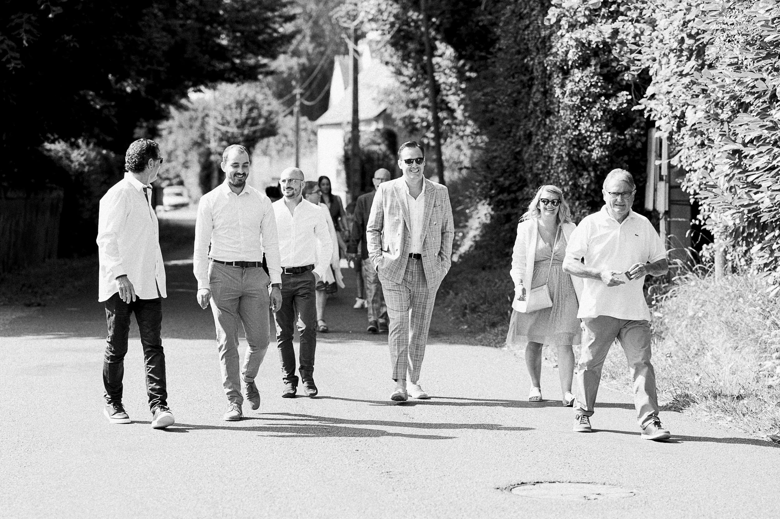 Groupe de personnes marchant ensemble sur un chemin en plein air, certaines portant des lunettes de soleil, dans un environnement verdoyant.