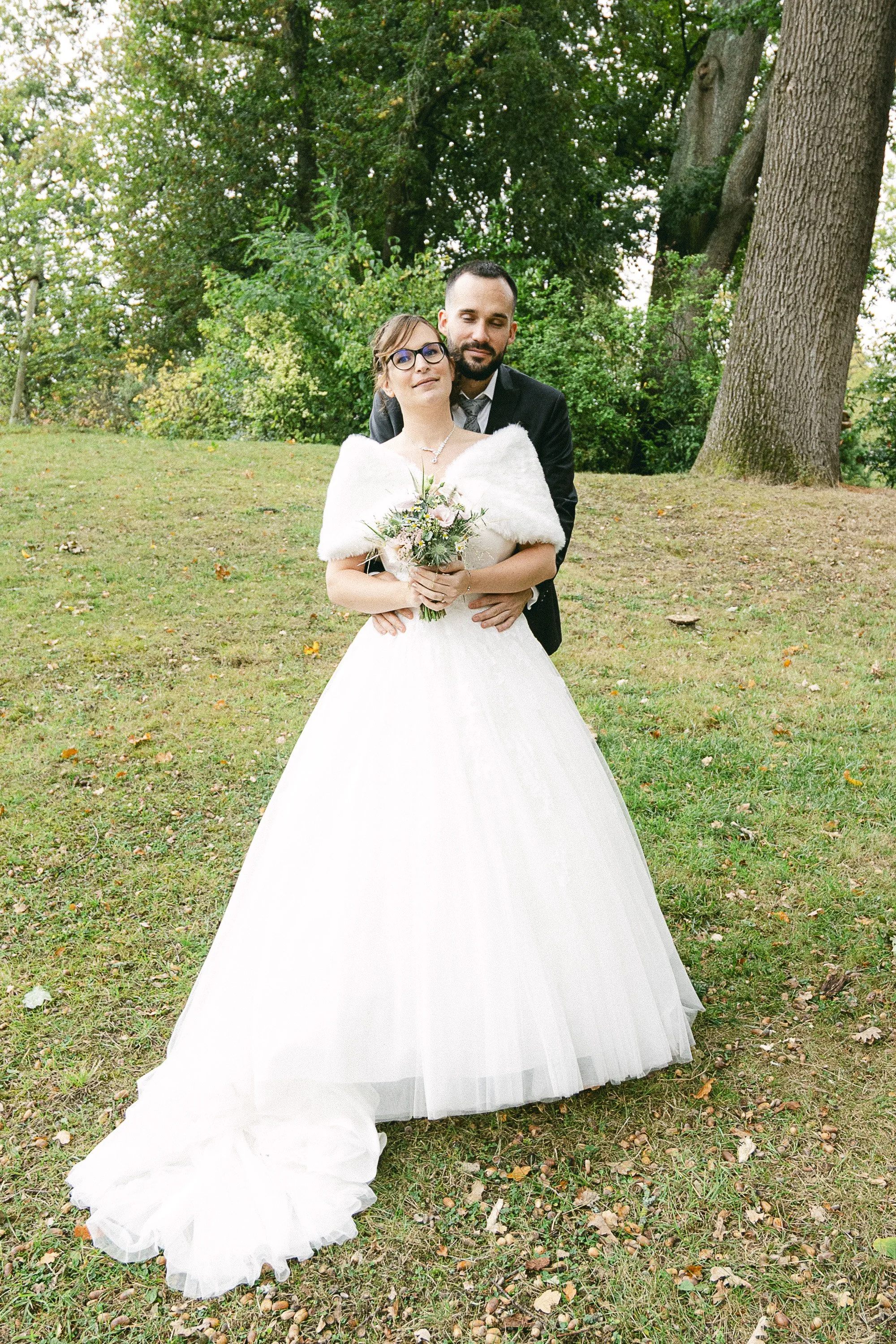 Un couple lors d'un mariage en plein air, la femme porte une robe blanche longue avec un manteau blanc et tient un bouquet, l'homme porte un costume noir.