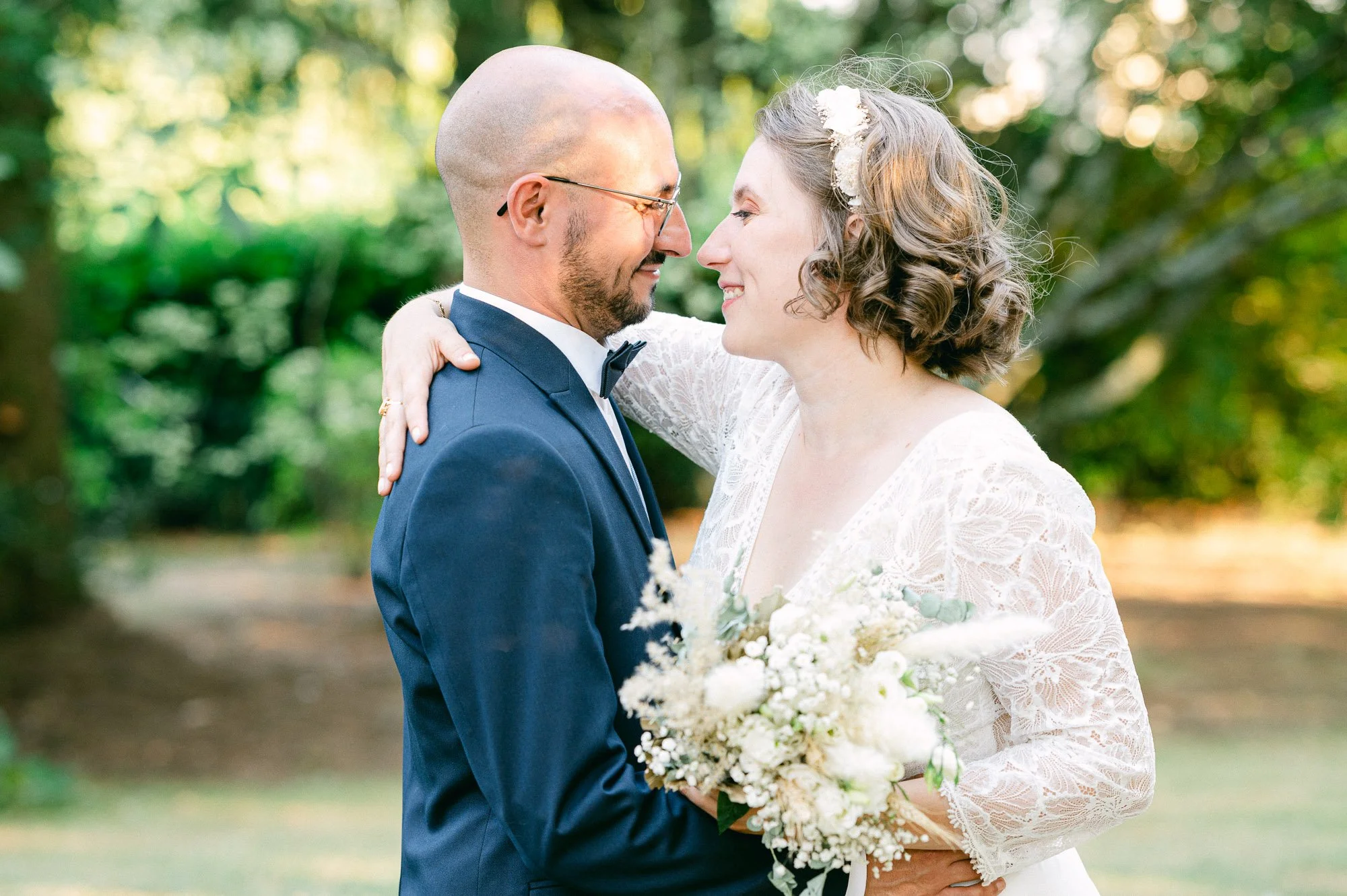Un couple de mariés dans un parc, se regardant tendrement, avec la femme tenant un bouquet de fleurs blanches.
