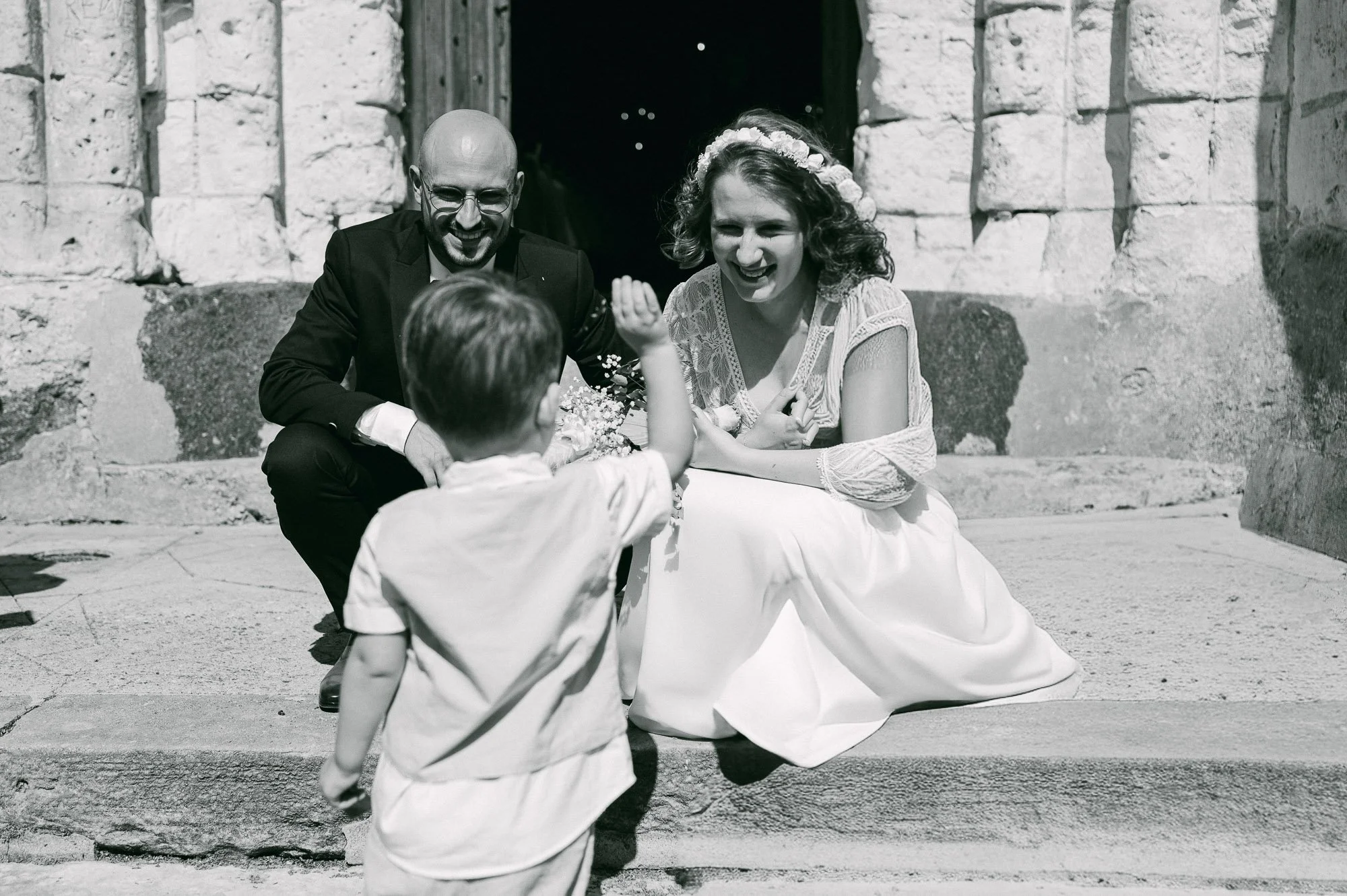 Une scène en noir et blanc d'un mariage où un couple de mariés rit avec deux jeunes enfants. La mariée porte une couronne de fleurs et une robe blanche, le marié porte un costume noir. Les enfants jouent et interagissent avec les mariés.