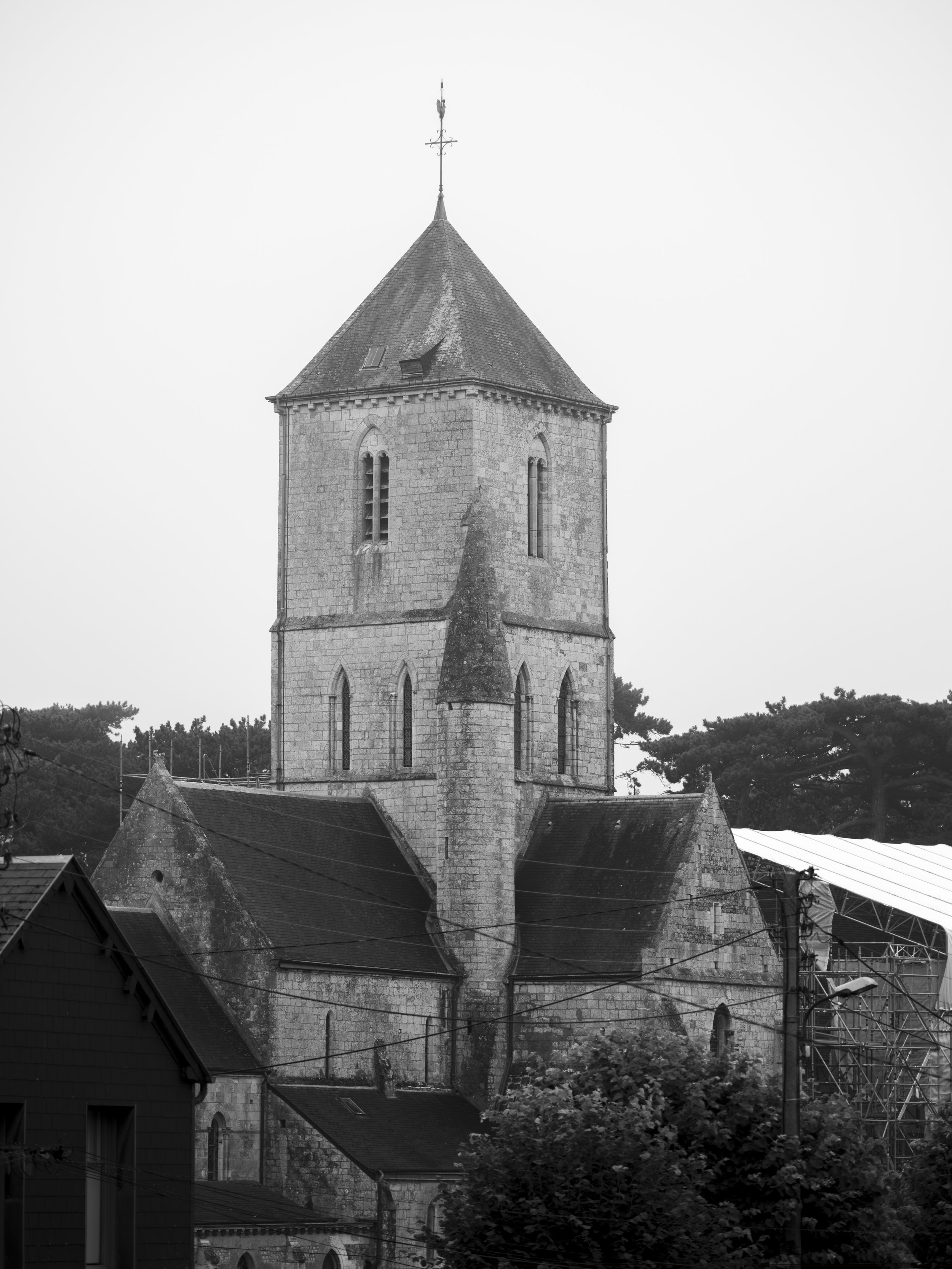Une vieille tour d'église en pierre avec un toit en pente, entourée de bâtiments et de végétation, en noir et blanc (Etretat)