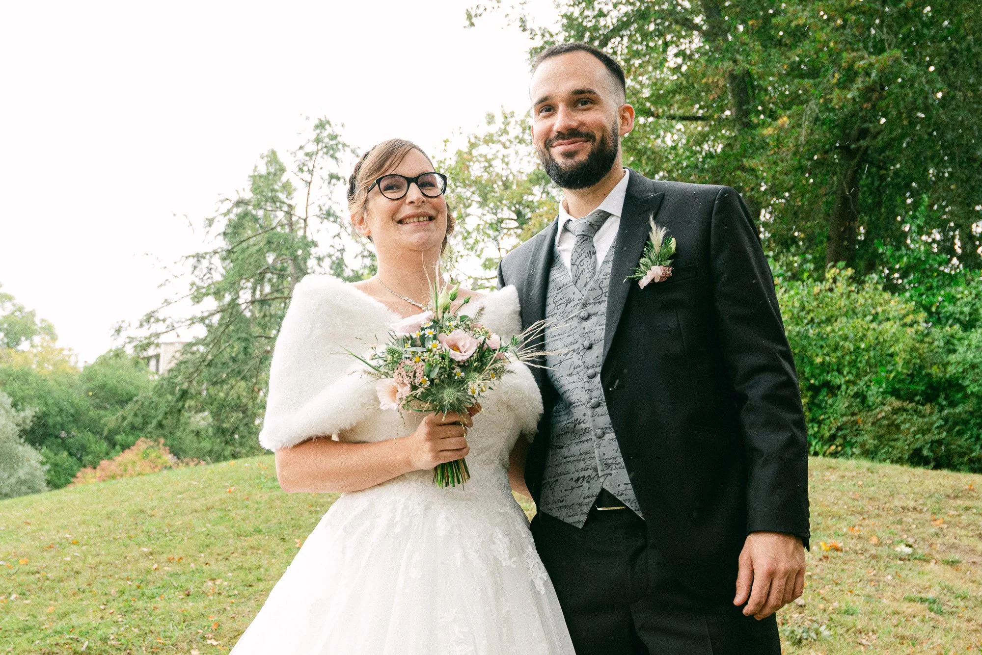 Un couple de mariés en robe de mariage et costume, souriants et posant dans un parc sur fond d'arbres verts, lors d'une journée en plein air.