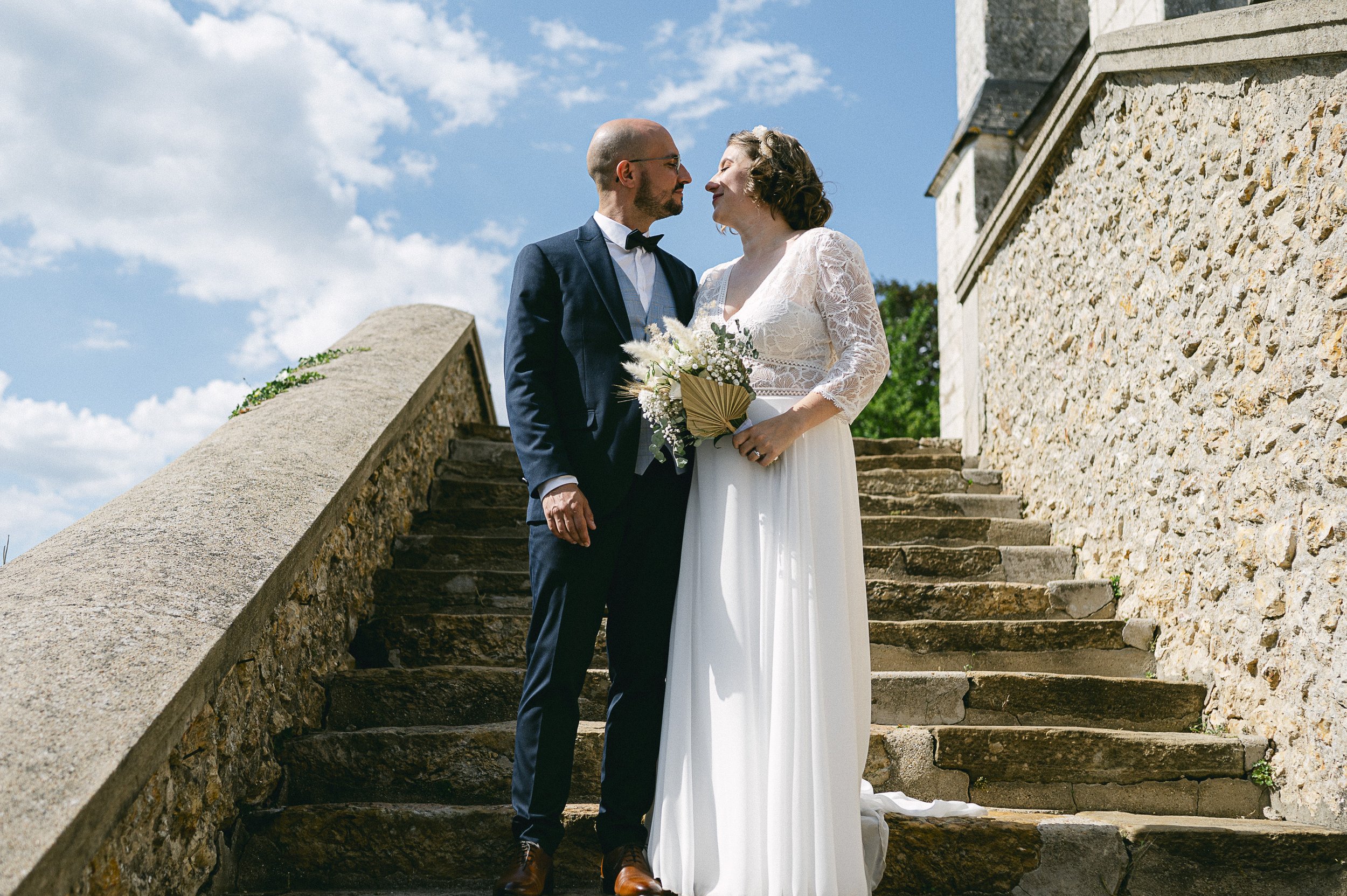 Un couple de mariés, homme en costume sombre et femme en robe blanche, se tiennent face à face en haut d'escaliers en pierre, avec un ciel bleu et quelques nuages en arrière-plan.