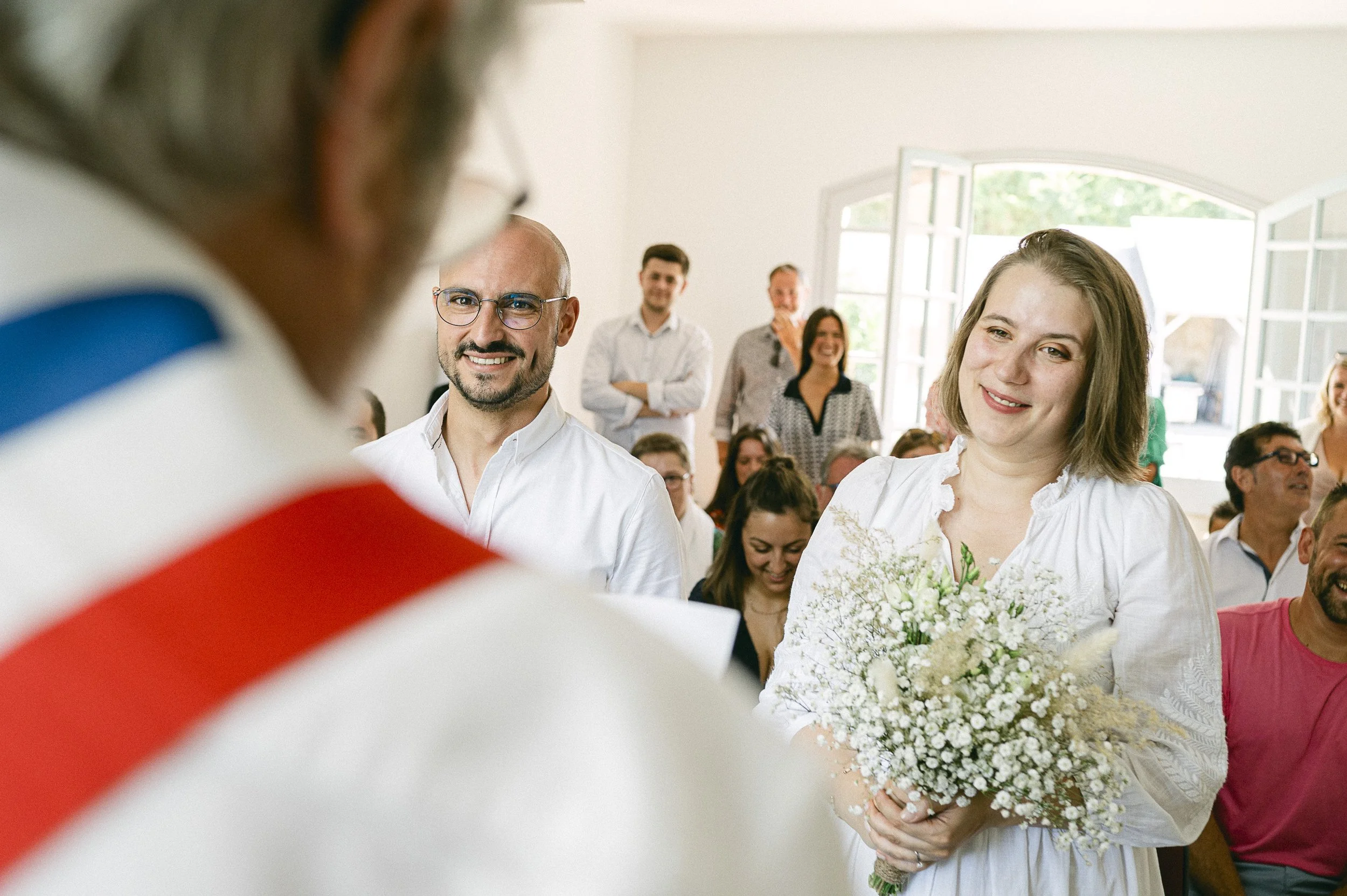 Une femme souriante tenant un bouquet de fleurs, lors d'une cérémonie ou d'un événement, entourée de plusieurs personnes dans une pièce lumineuse.