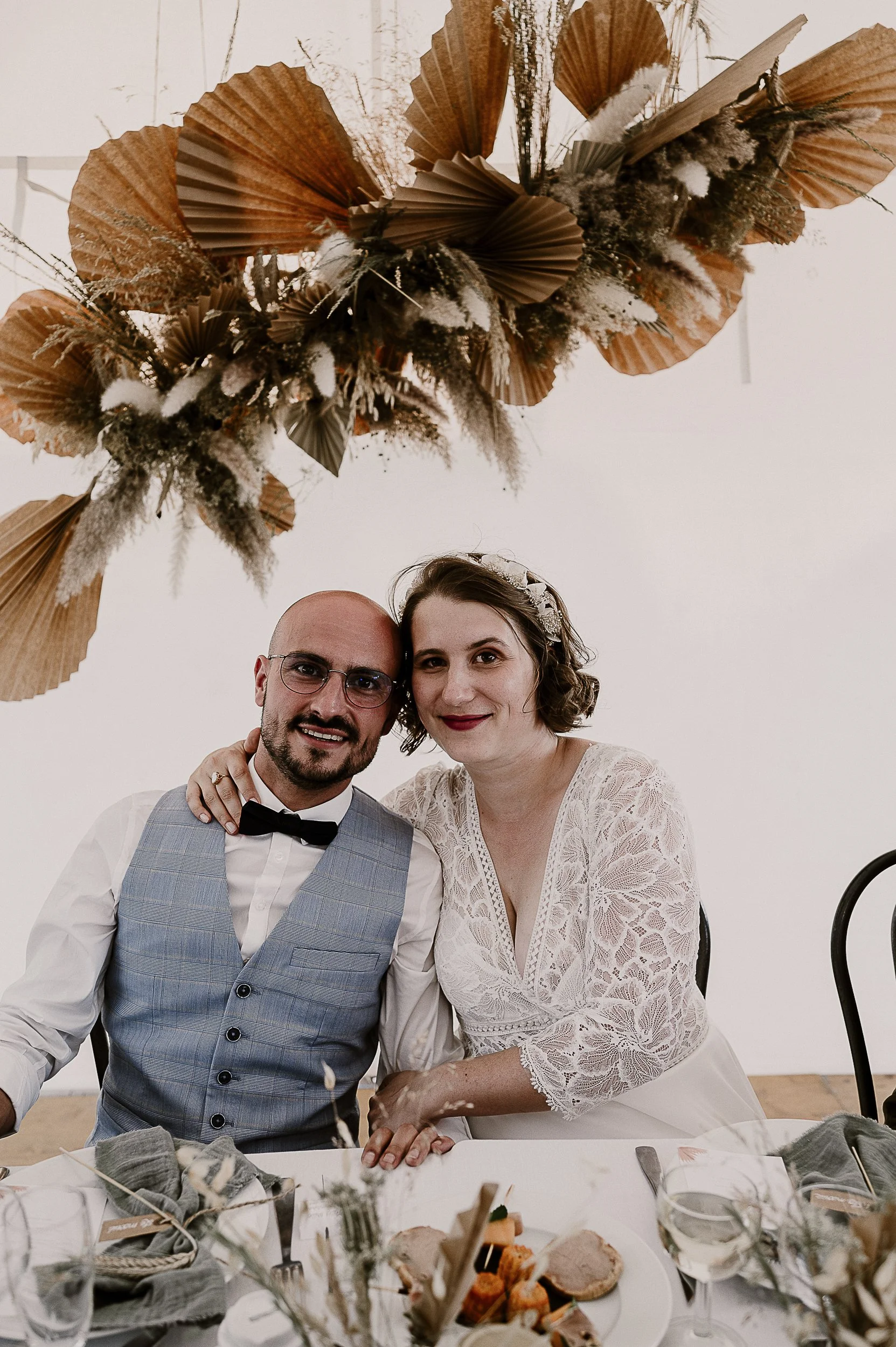 Un couple lors d'une célébration, probablement un mariage, assis à une table décorée avec de la vaisselle et des mets, avec une décoration florale suspendue au plafond.