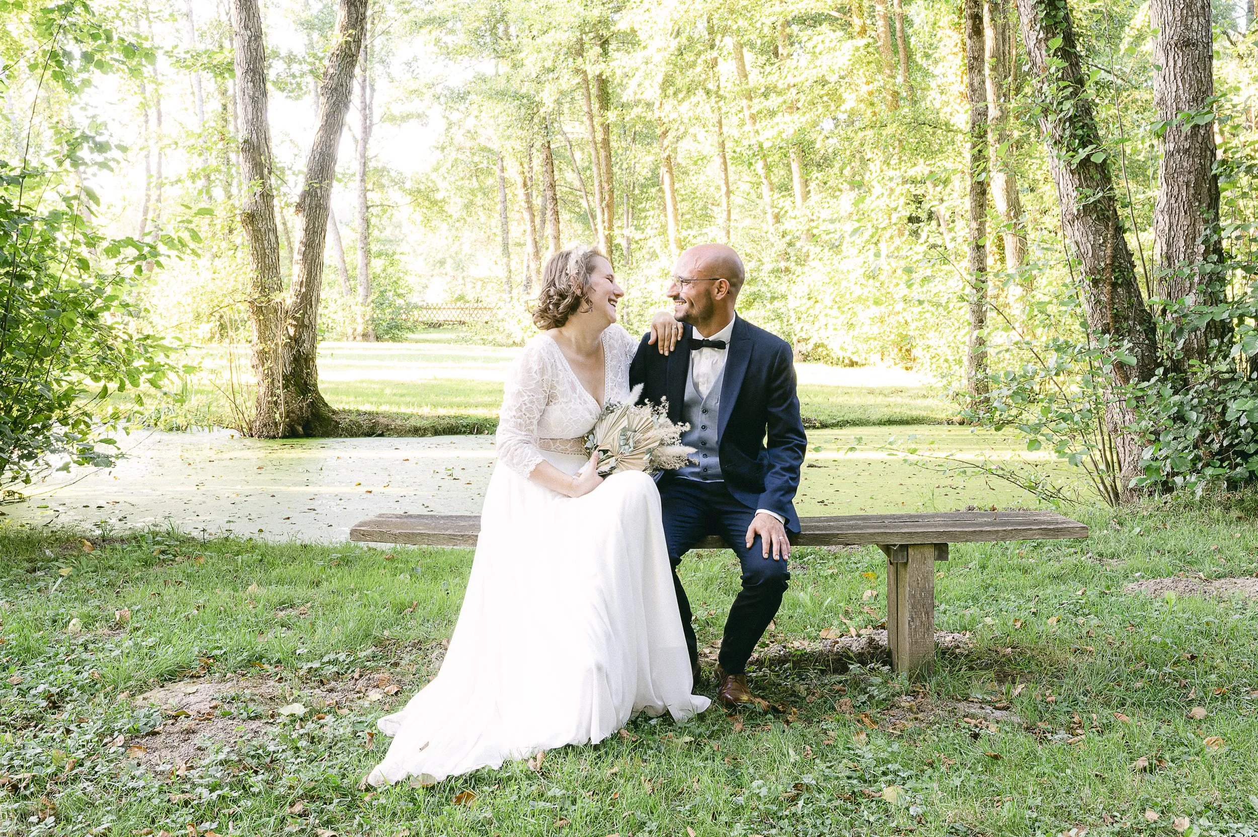 Un couple vêtu de vêtements de mariage, assis sur un banc en bois dans une forêt verdoyante, partageant un moment joyeux et complice, avec des arbres et des feuilles en arrière-plan.