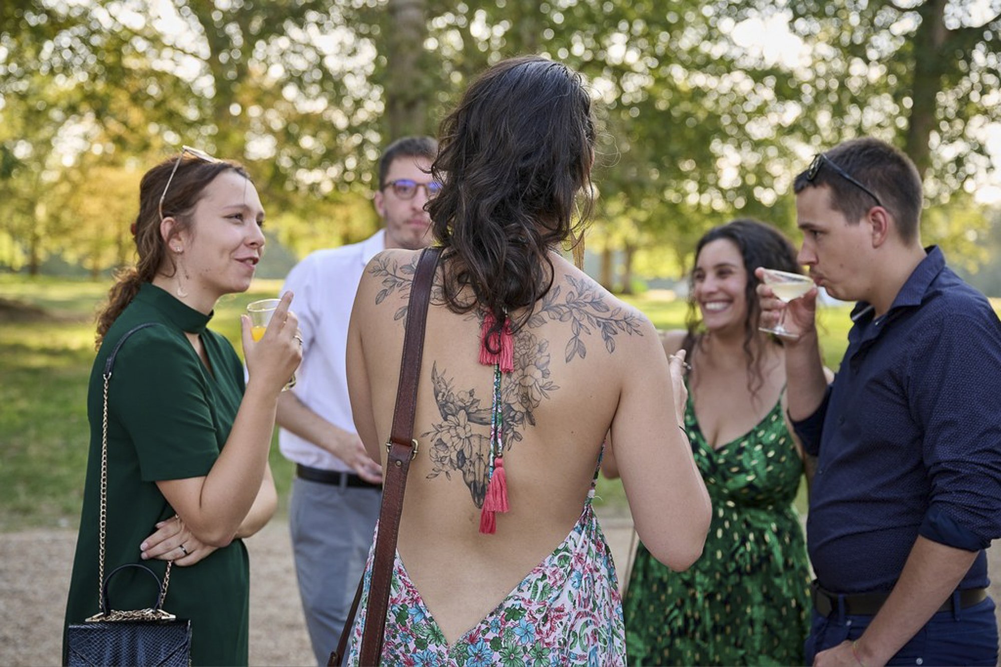 Un groupe de jeunes adultes discutant dans un parc lors d'une journée ensoleillée. La femme au centre a des tatouages dans le dos, porte une robe à fleurs, et tient une boisson. Les autres sont habillés de manière décontractée, certains portent des l