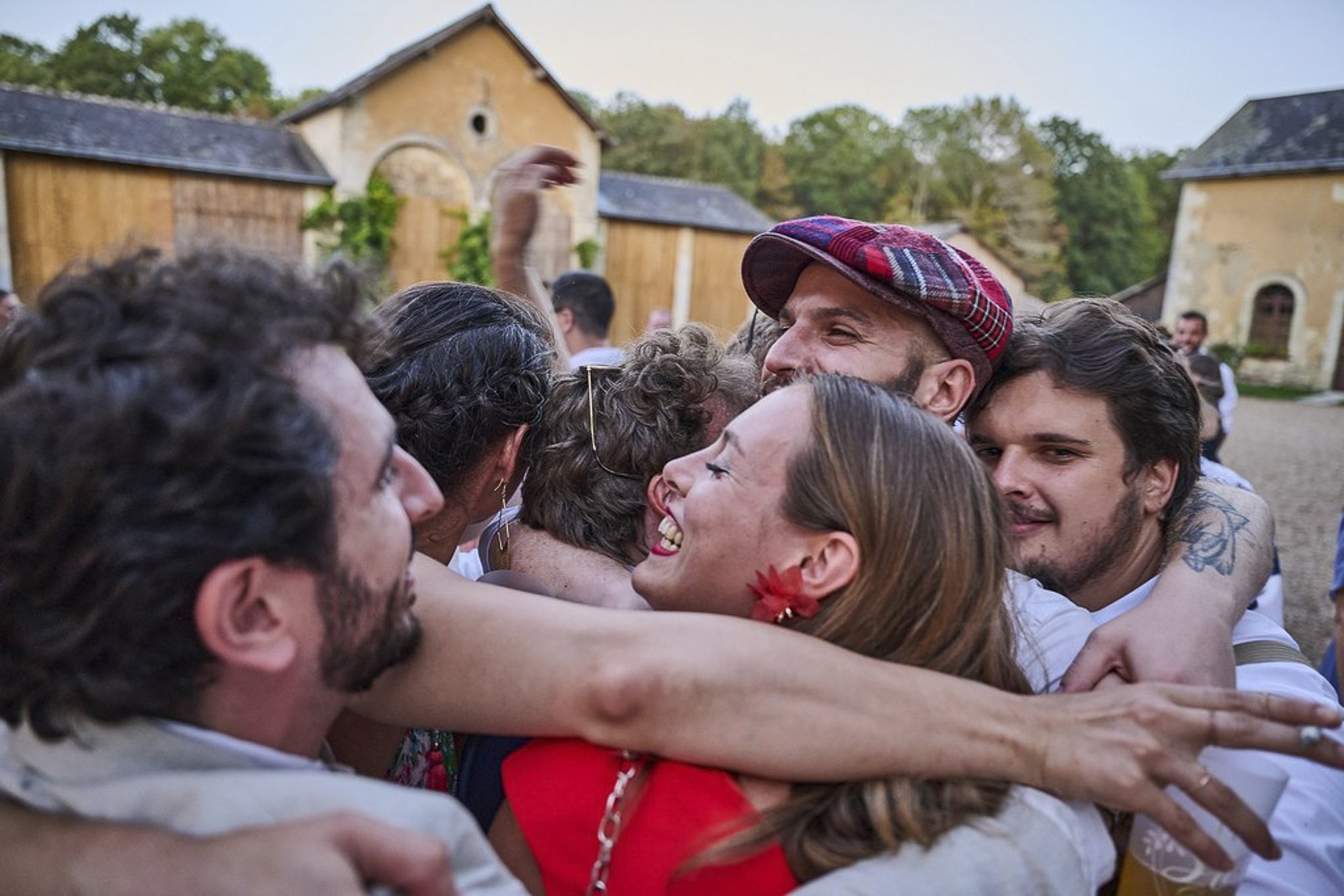 Groupe de personnes en costume d'été embrassant et souriant lors d'une célébration en extérieur devant des bâtiments en pierre avec des toits en tuiles.