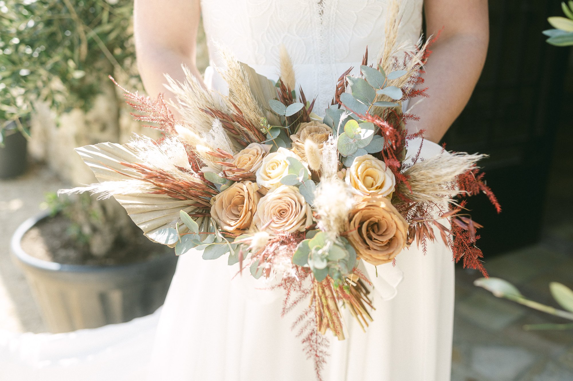 Un bouquet de fleurs avec des roses, des feuilles d'eucalyptus et des plantes séchées, porté par une personne en robe blanche, lors d'une cérémonie ou d'un mariage en extérieur.