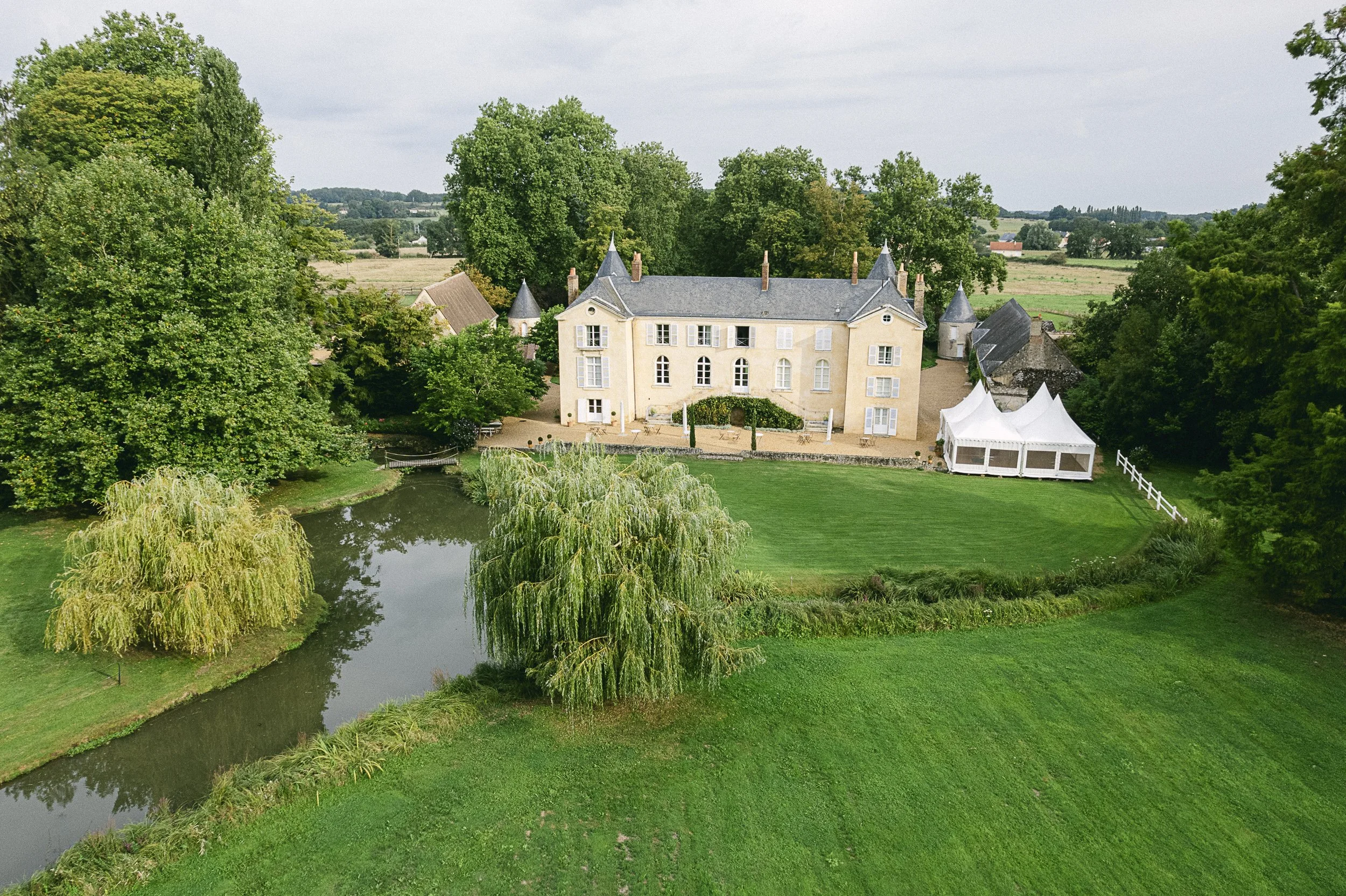 Un manoir ancienne avec un toit en ardoise, entouré d'arbres verts et d'un petit étang avec des cerisiers en fleurs à proximité, dans un paysage rural paisible.