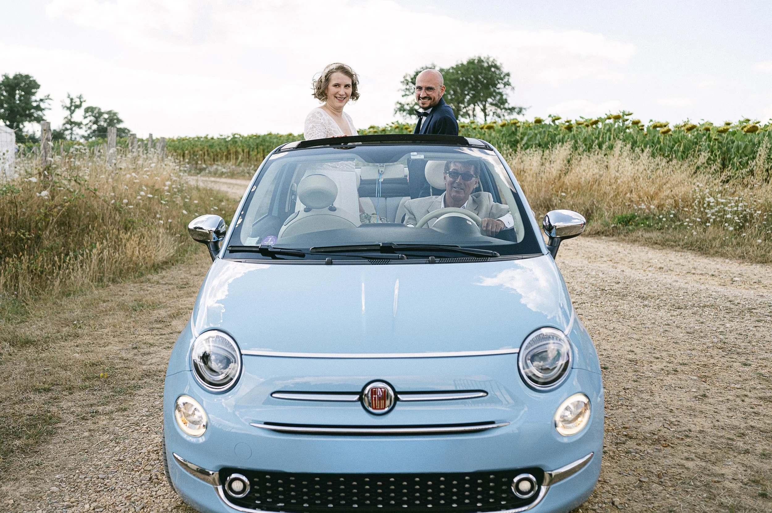 Deux personnes, une femme en robe blanche et un homme en costume, debout dans une voiture décapotable bleue sur un chemin de campagne ensoleillé avec un champ de tournesols à l'arrière.