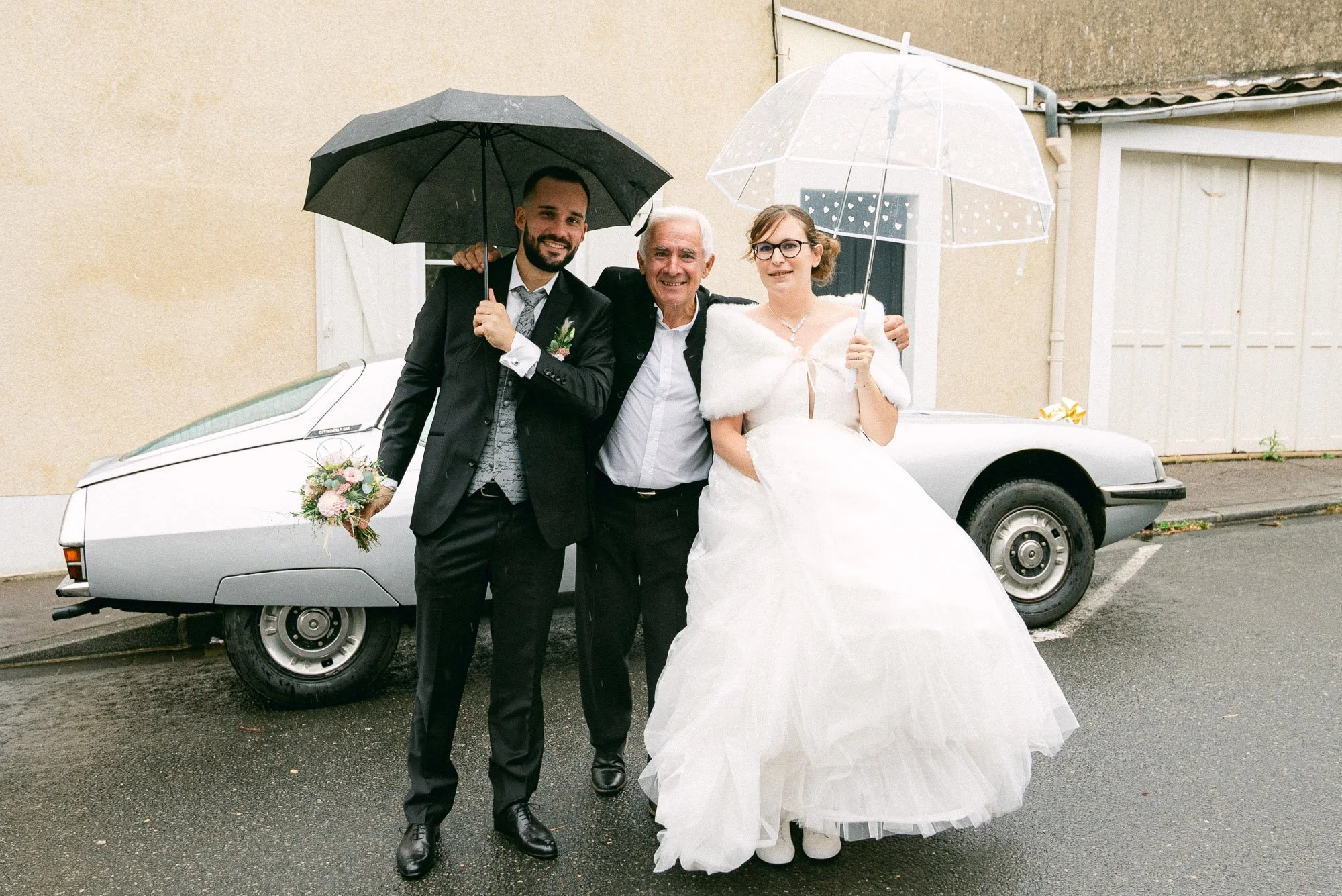 Trois personnes vêtues de vêtements de mariage sous des parapluies devant une voiture blanche, en train de célébrer un mariage.