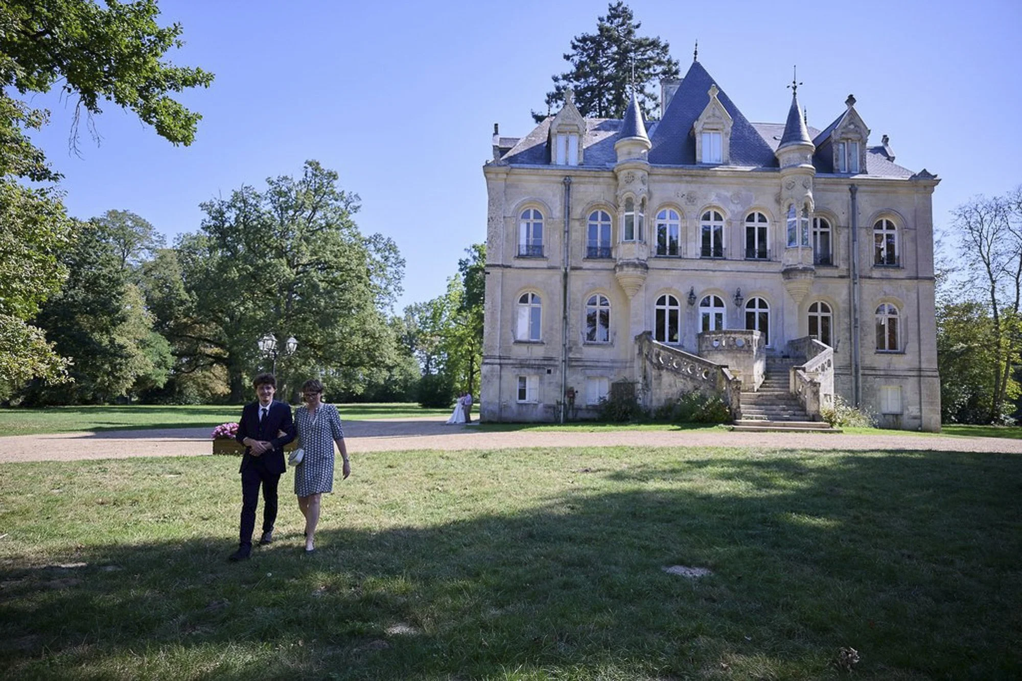 Un château victorien en pierre avec plusieurs tourelles et fenêtres, entouré d'arbres verts, avec deux personnes en vêtements formels marchant sur la pelouse en premier plan.
