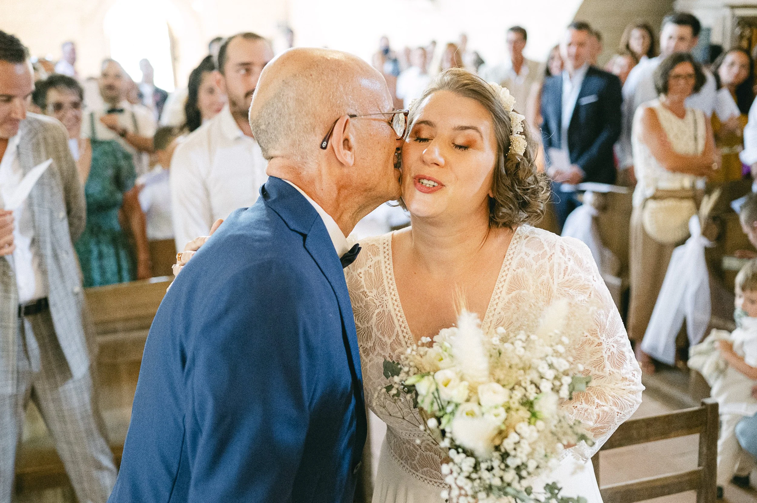 Un couple marié échange un baiser lors d'une cérémonie religieuse ou civile, entouré d'invités assis et debout.