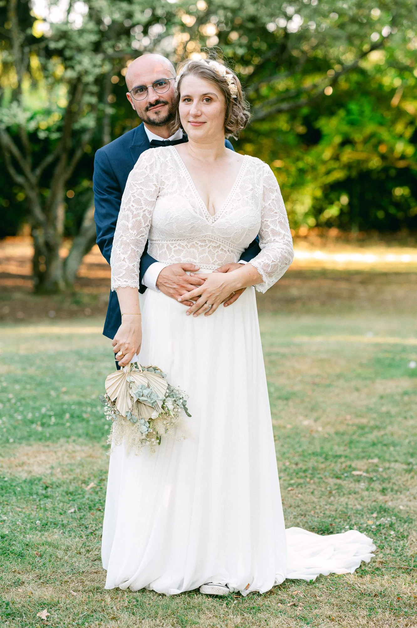 Un couple de mariage posant dans un parc, la femme en robe blanche et le homme en costume bleu marine avec noeud papillon, tenant un bouquet de fleurs.