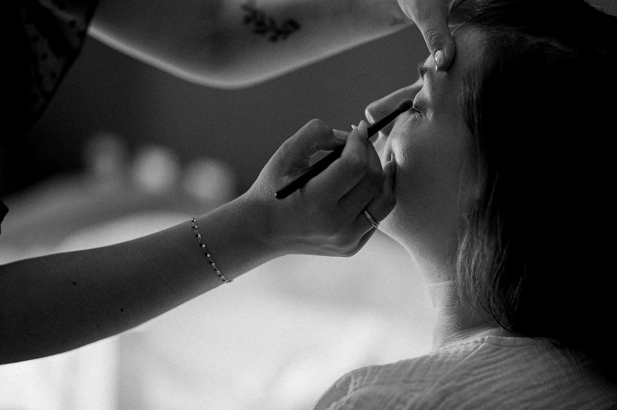 Une image en noir et blanc montrant une femme en train de se faire maquiller l'œil, avec un pinceau. La scène est prise de profil, et une main tient le pinceau près de son œil.