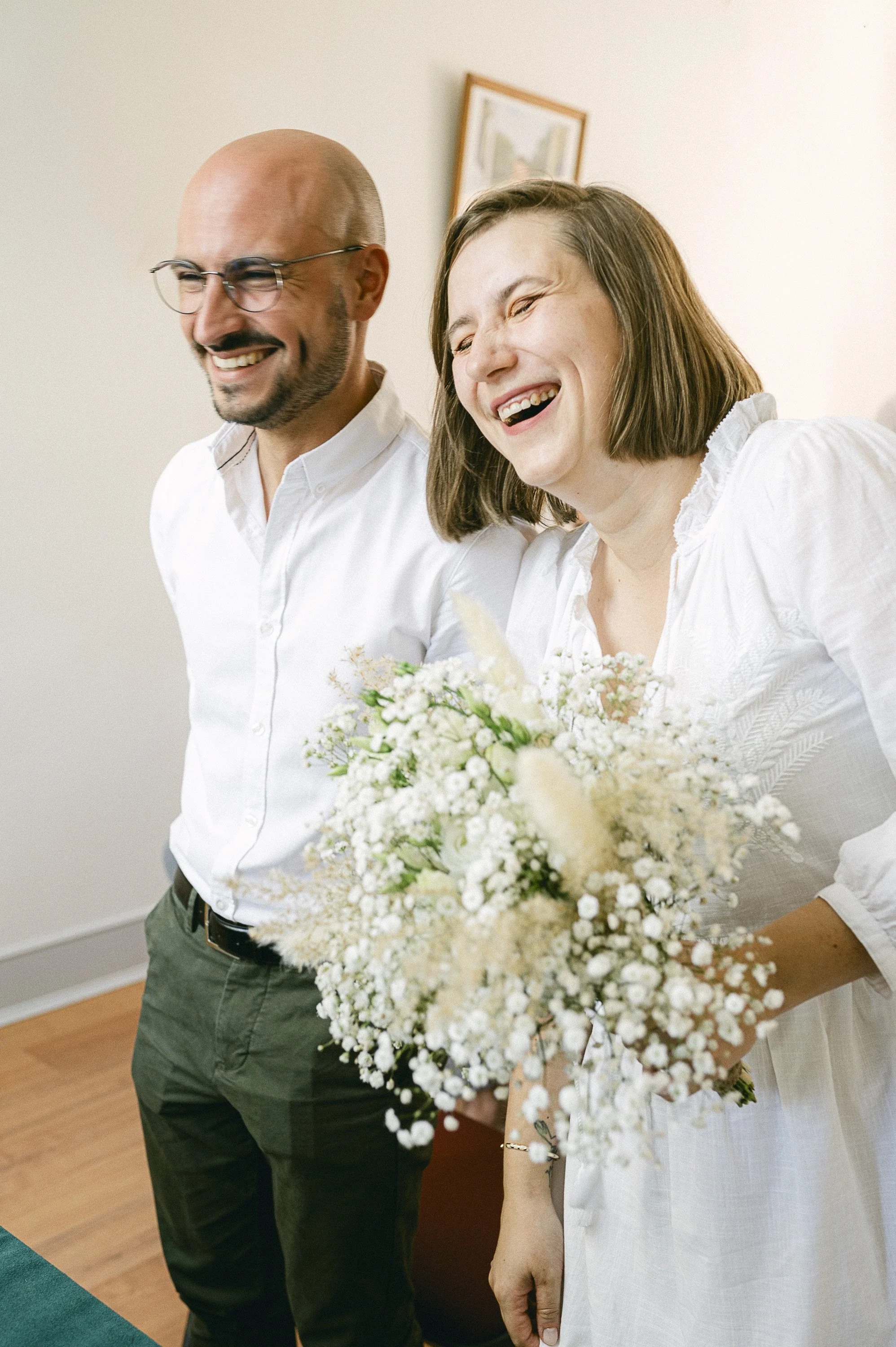 Deux personnes, un homme et une femme, sourient lors d'une occasion spéciale, la femme tient un bouquet de fleurs blanches.