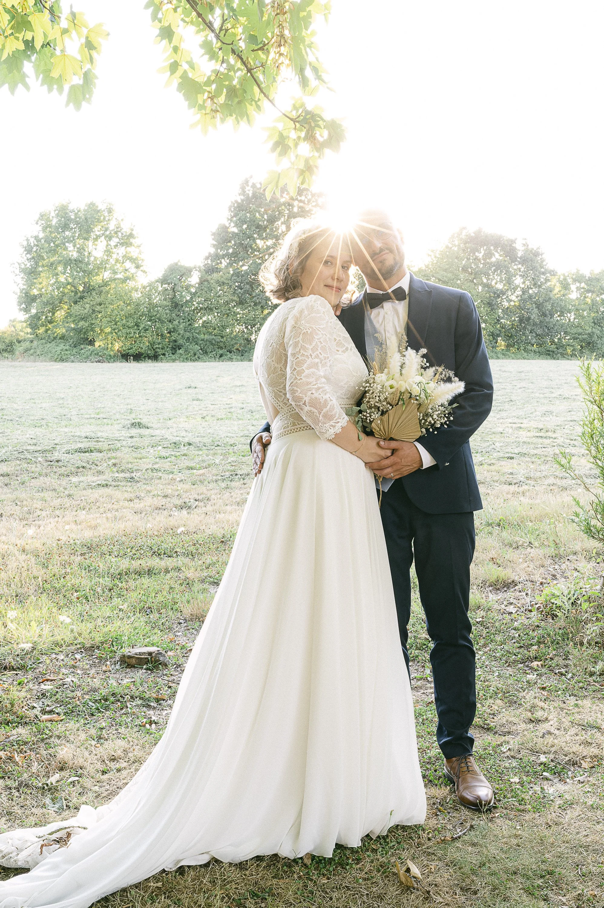 Un couple de mariage pose dans un champ en plein air, la femme porte une robe de mariée blanche avec un bouquet de fleurs et l'homme porte un costume noir avec un nœud papillon. Le soleil brille derrière eux, créant un effet de halo.