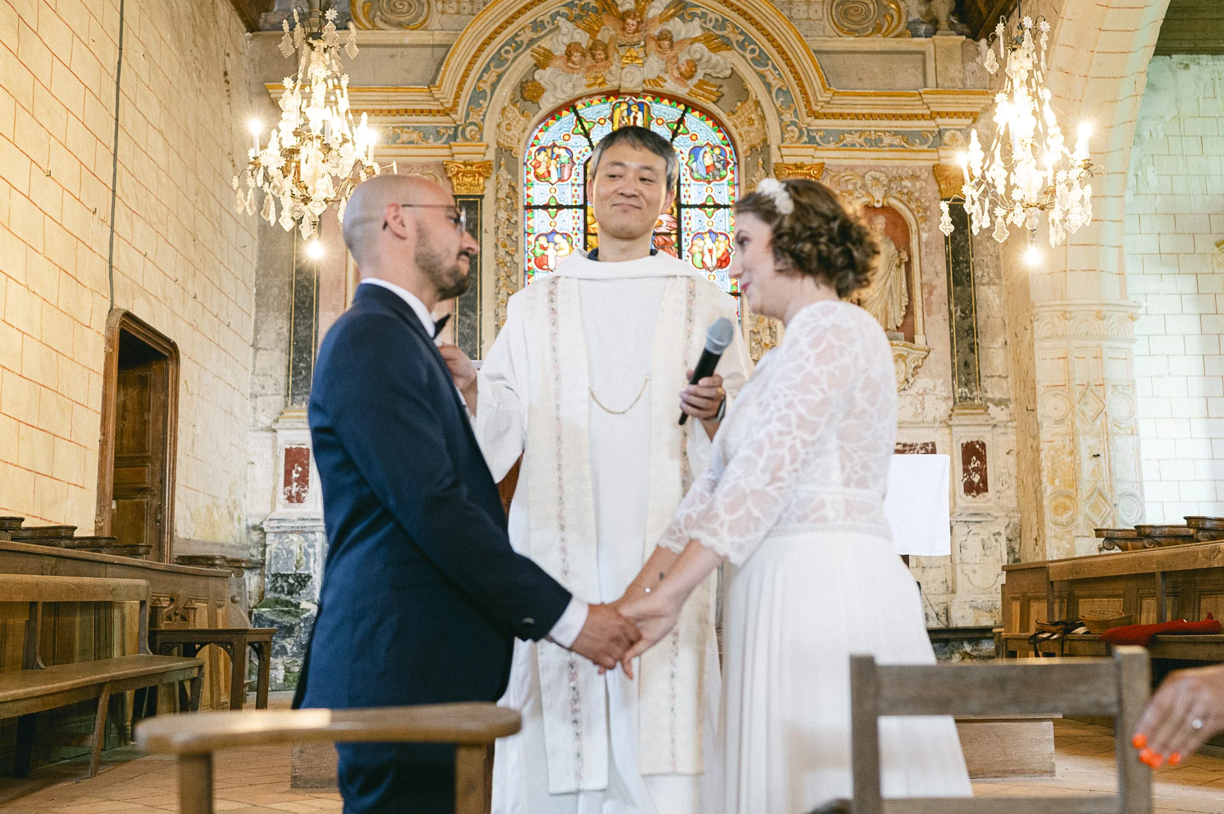 Un couple de mariés lors de leur cérémonie de mariage religieux dans une église, avec un prêtre au centre. Les mariés se tiennent par la main, dans un décor d'église avec des vitraux colorés et des chandeliers