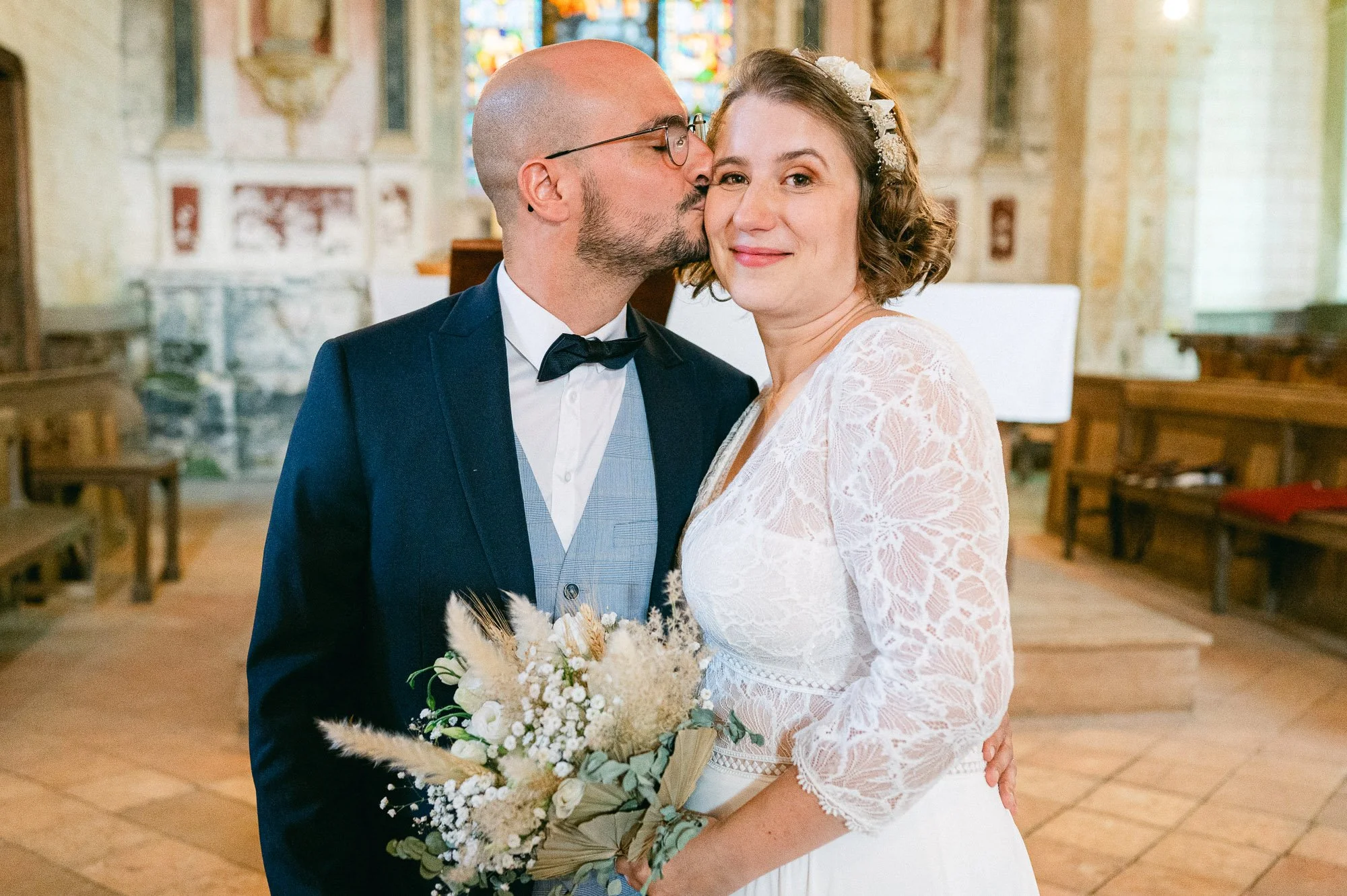 Un couple de mariés dans une église, l'homme donne un bisou sur la joue de la femme, qui tient un bouquet de fleurs. L'intérieur de l'église est décoré avec des vitraux colorés en arrière-plan.