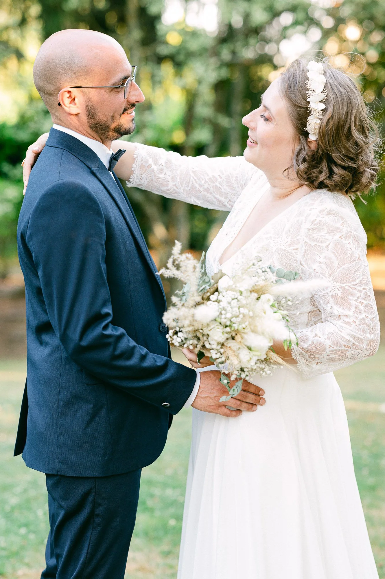 Un couple marié échangeant un baiser lors d'une cérémonie en plein air, la femme portant une robe blanche et un bandeau floral dans les cheveux, tenant un bouquet de fleurs blanches. L'homme porte un costume bleu marine avec une cravate noire.
