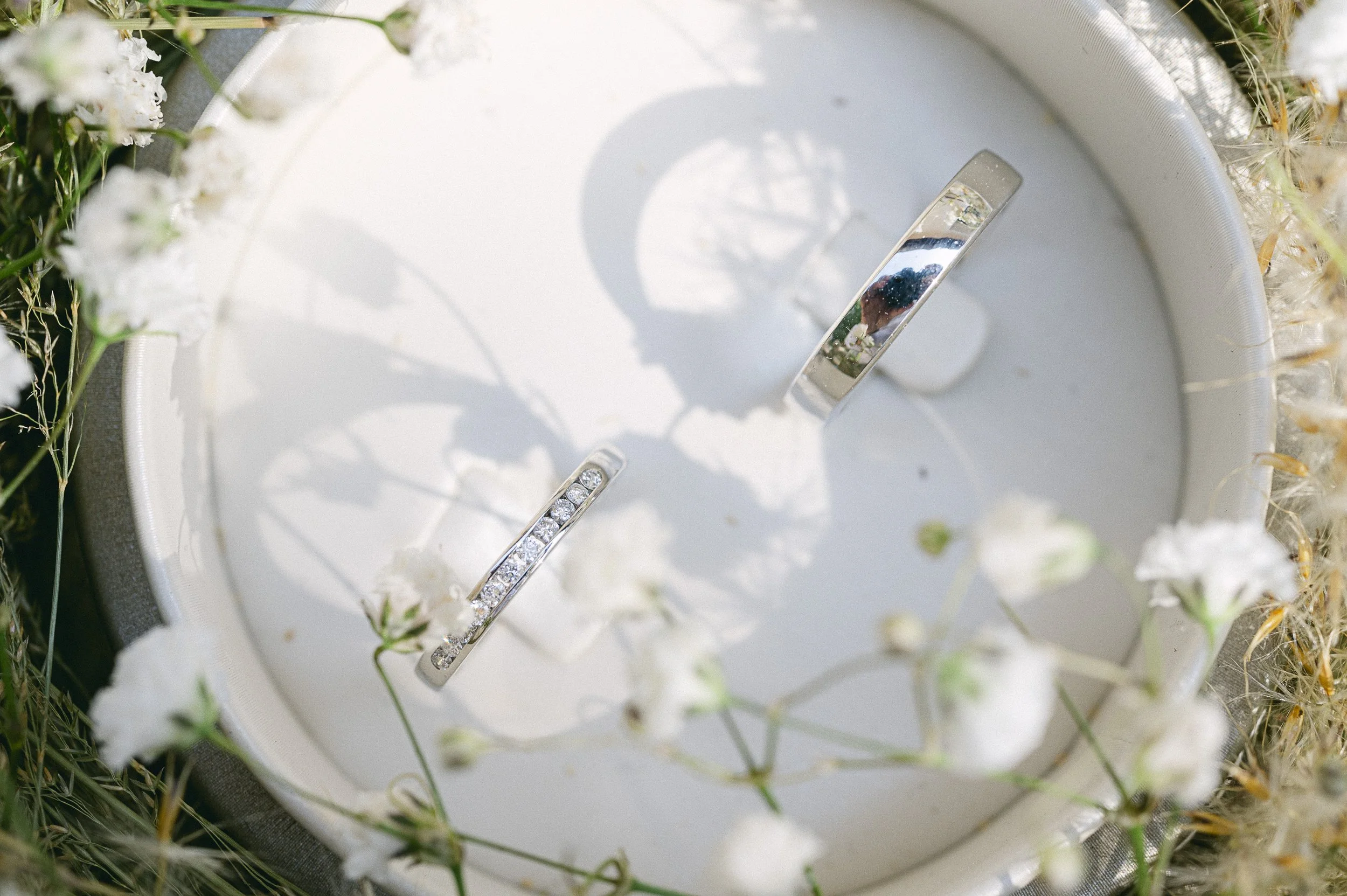 Anneaux de mariage posés sur un coussin blanc entouré de fleurs blanches et de petites plantes. La photo est prise en extérieur, avec un reflet visible dans un anneau.