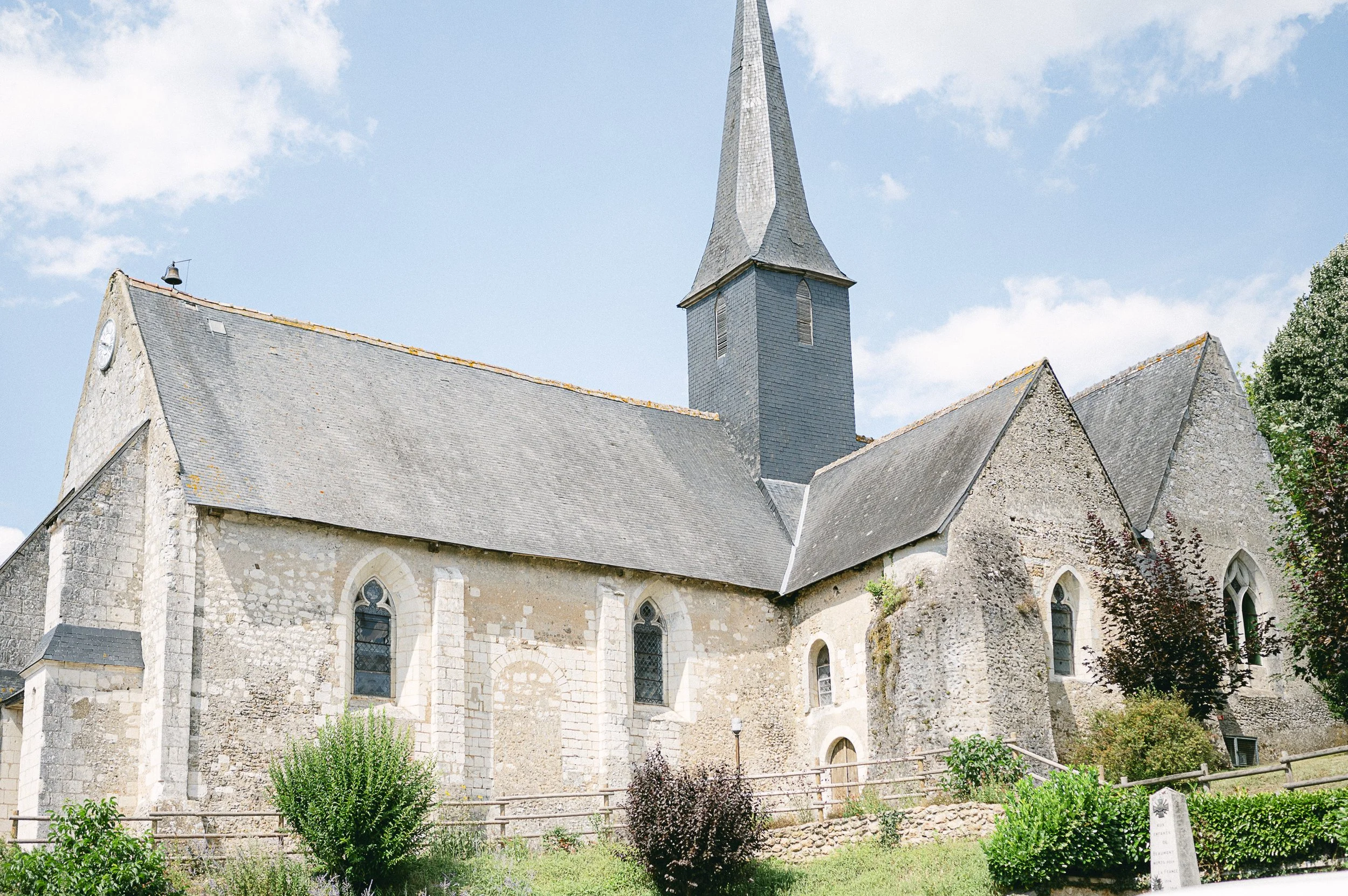 Église en pierre avec un toit en ardoise et une grande tour avec un clocher en pierre, entourée de végétation verte.