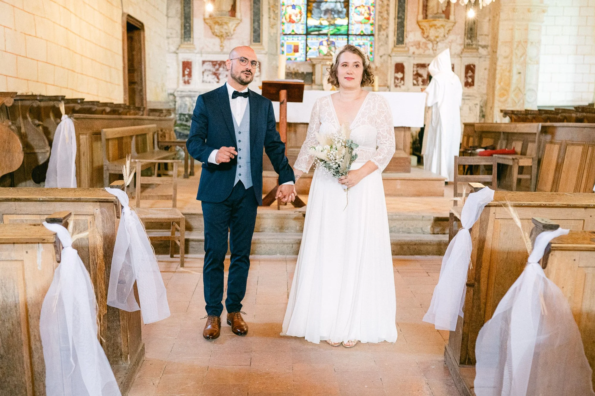 Un couple lors d'un mariage dans une église, la femme en robe blanche tenant un bouquet de fleurs, l'homme en costume sombre avec nœud papillon, entourés de bancs décorés de rubans blancs, dans une église lumineuse avec vitraux colorés.