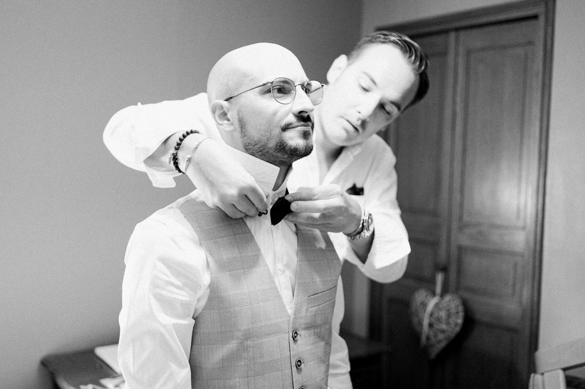 Deux hommes en costume blanc et gilet, l’un portant des lunettes, se montrent mutuellement dans une salle, en noir et blanc.