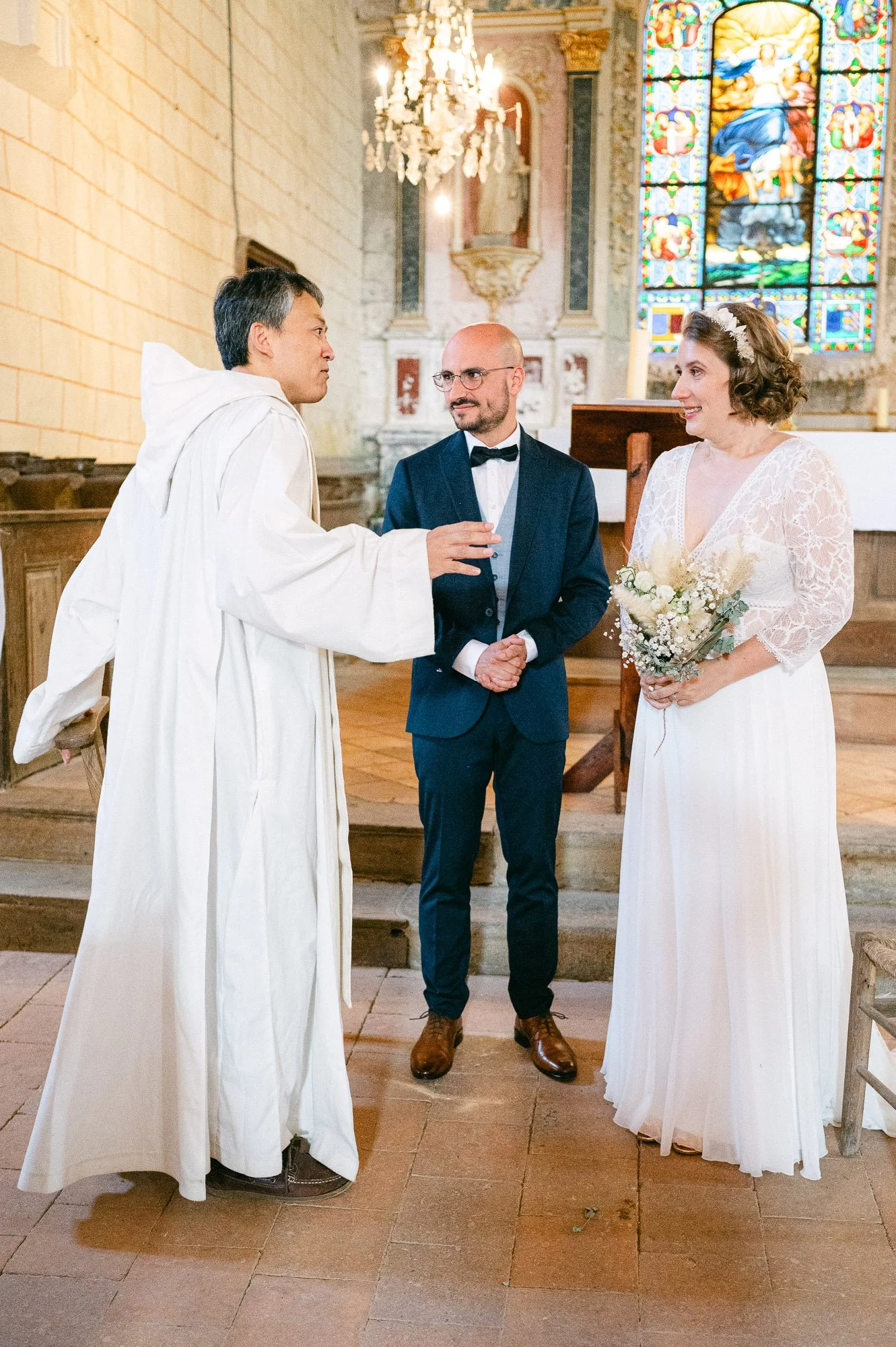 Un couple se marie dans une église, avec un prêtre en costume bleu et une femme en robe blanche tenant un bouquet de fleurs. L'intérieur de l'église est orné d'une grande fenêtre en vitrail coloré et d'un chandelier chandelier suspendu au plafond.
