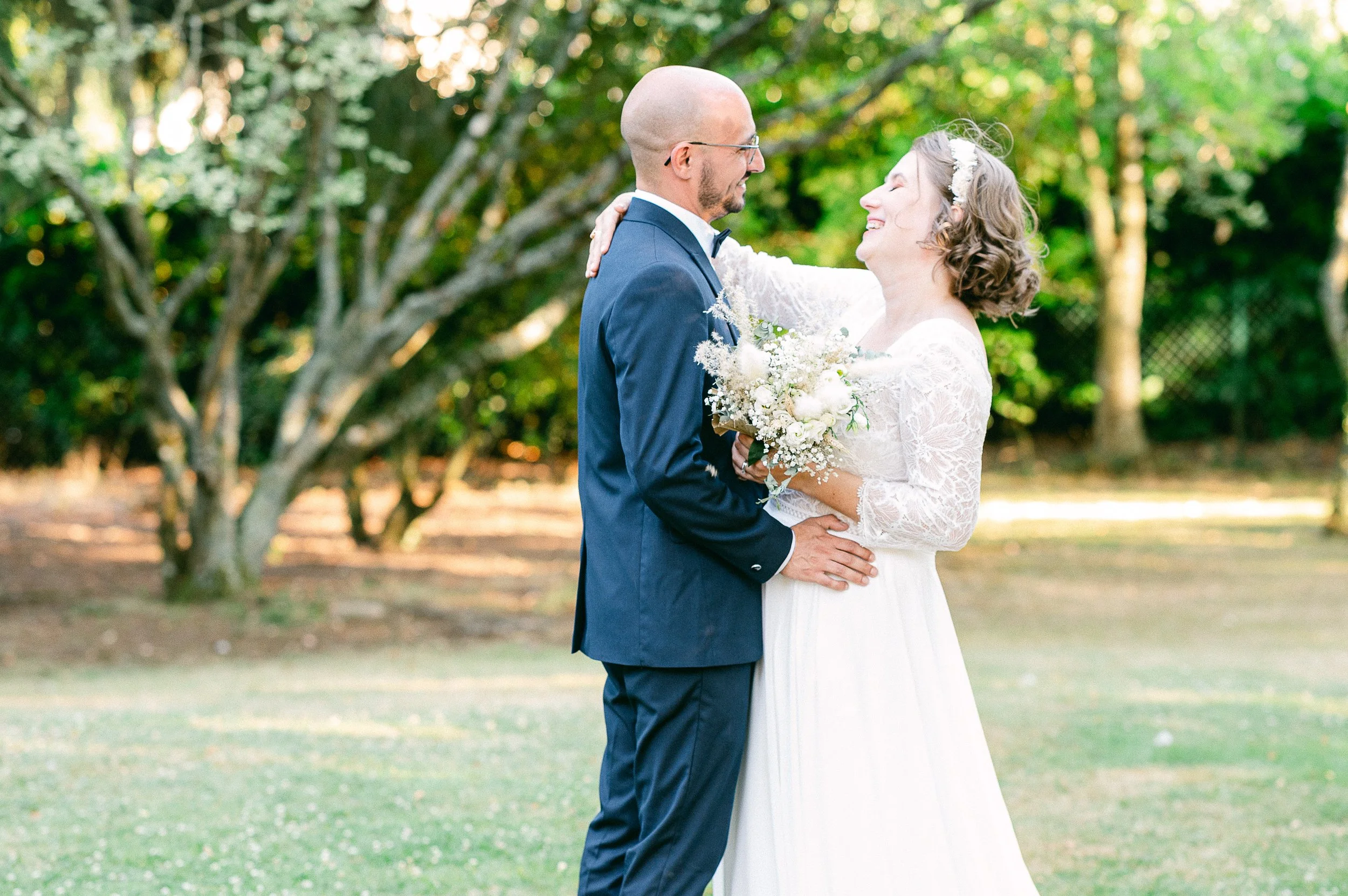 Un couple en robe de mariage et costume debout dans un parc, se regardant avec bonheur, entouré d'arbres verts.