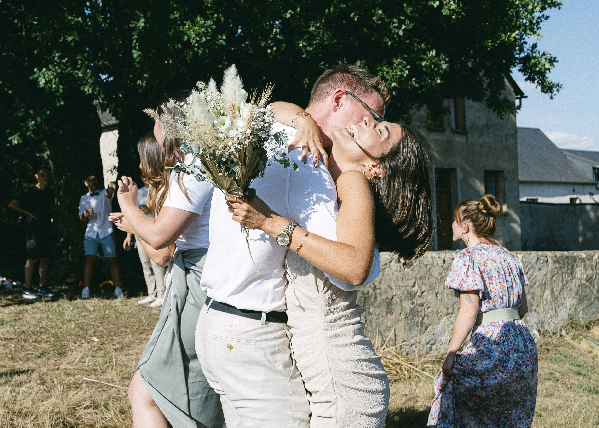 Deux personnes dansent et s'embrassent lors d'une fête en plein air, un homme tient un bouquet de fleurs tandis que la femme sourit avec les yeux fermés.