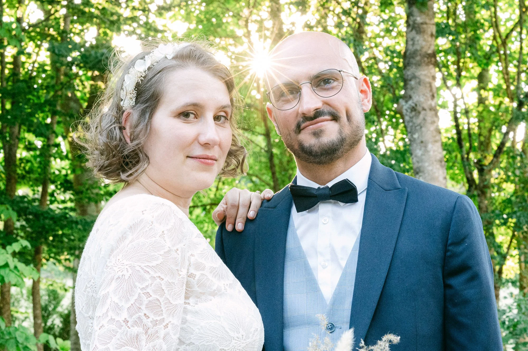 Un couple lors d'un mariage, la femme porte une robe blanche et un bandeau floral, l'homme porte un costume bleu avec nœud papillon. Ils se tiennent dans une forêt ensoleillée.