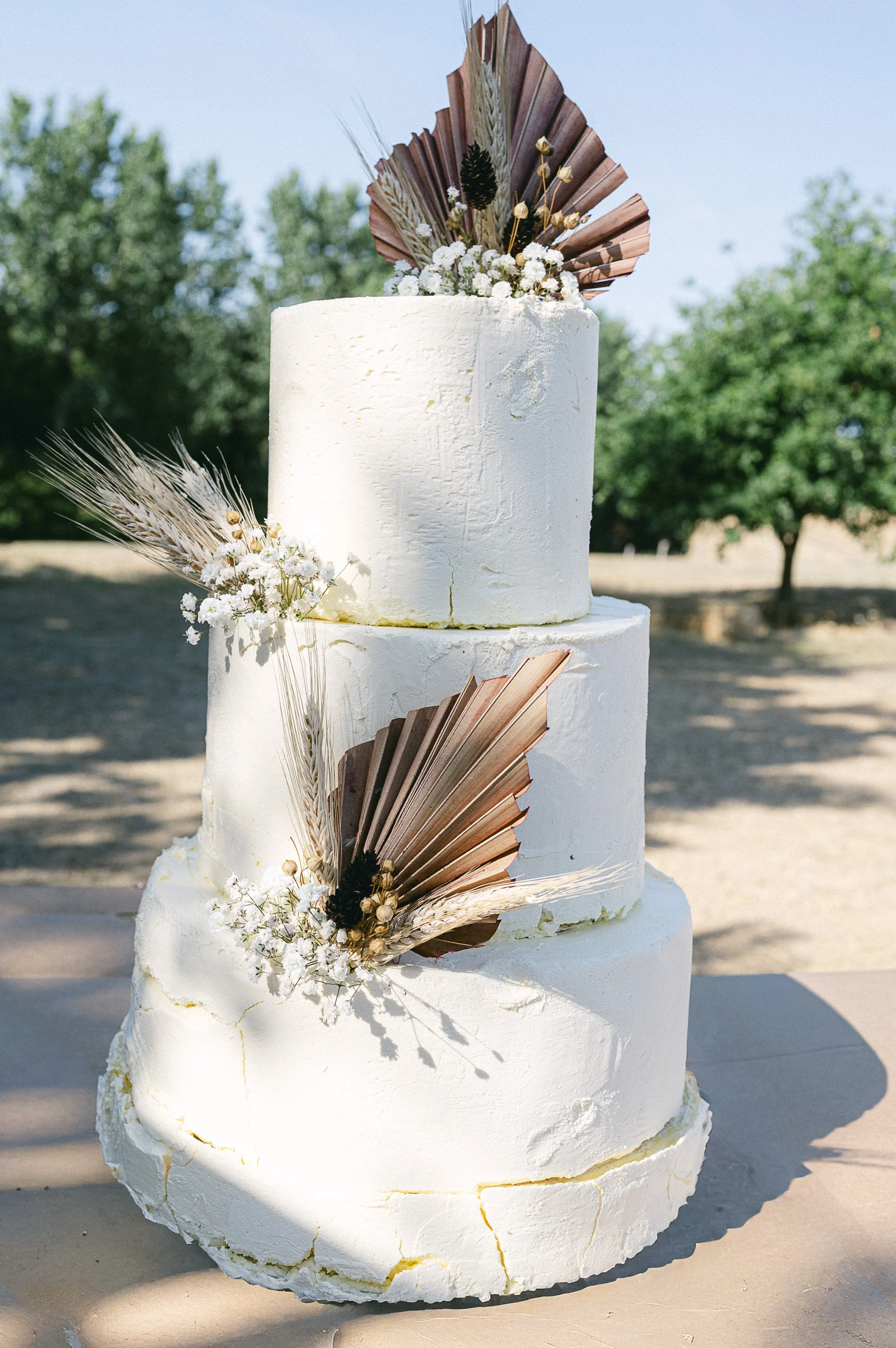 Gâteau de mariage à trois niveaux blanc orné de fleurs blanches et de feuilles en papier décoratives, placé en extérieur avec des arbres en fond.
