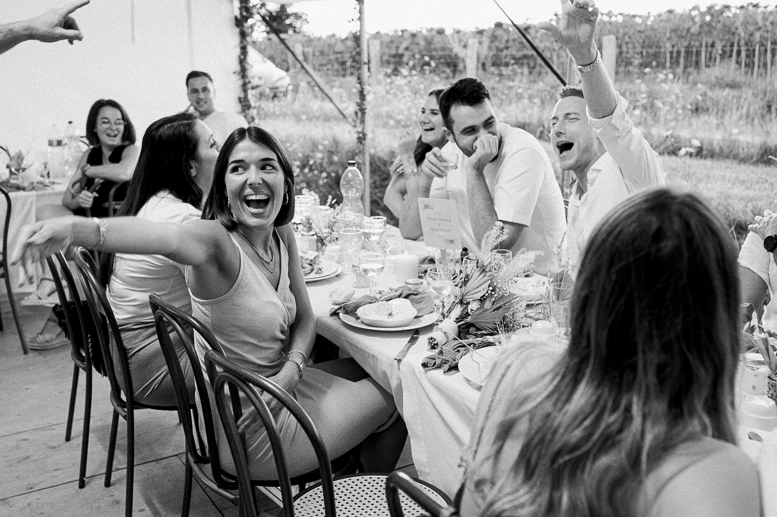 Groupe de personnes souriantes et riant lors d'un repas en plein air, autour d'une table avec de la nourriture et des boissons.