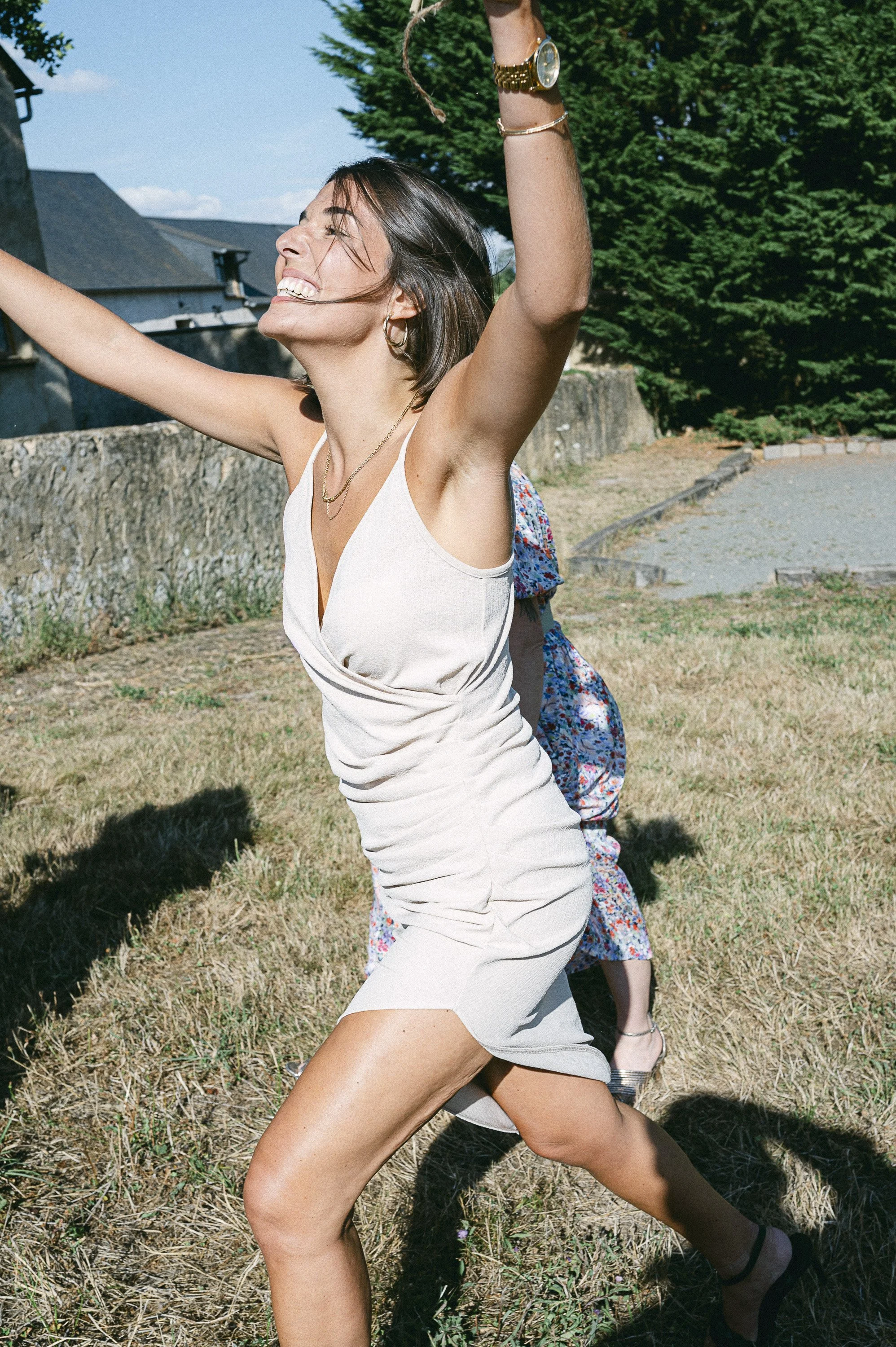 Une femme souriante et joyeuse avec les bras levés, portant une robe blanche, en train de danser ou de jouer dehors par une journée ensoleillée avec un arbre vert en arrière-plan.