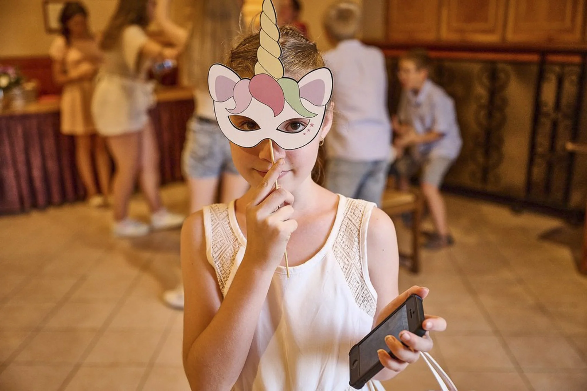 Une jeune fille porte un masque de licorne et tient un téléphone portable, en arrière-plan plusieurs personnes sont dans une pièce chaleureuse avec un décor en bois.