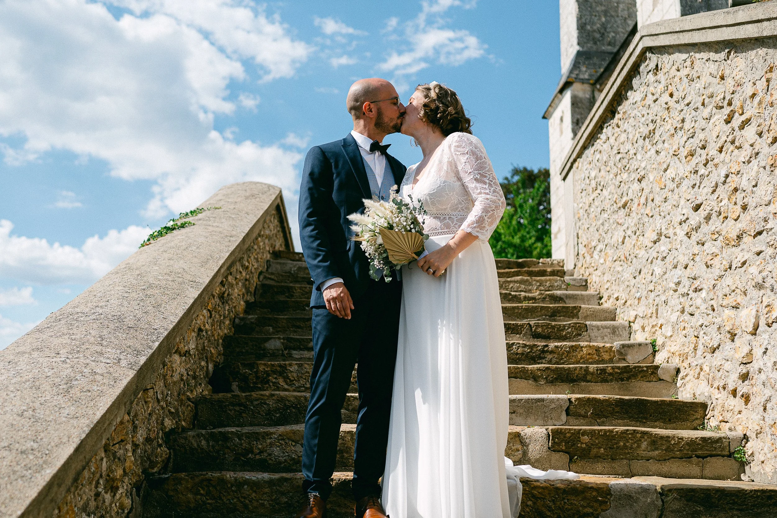 Un couple en mariage s'embrassant sur un escalier en pierre, avec un ciel bleu et des nuages en arrière-plan.