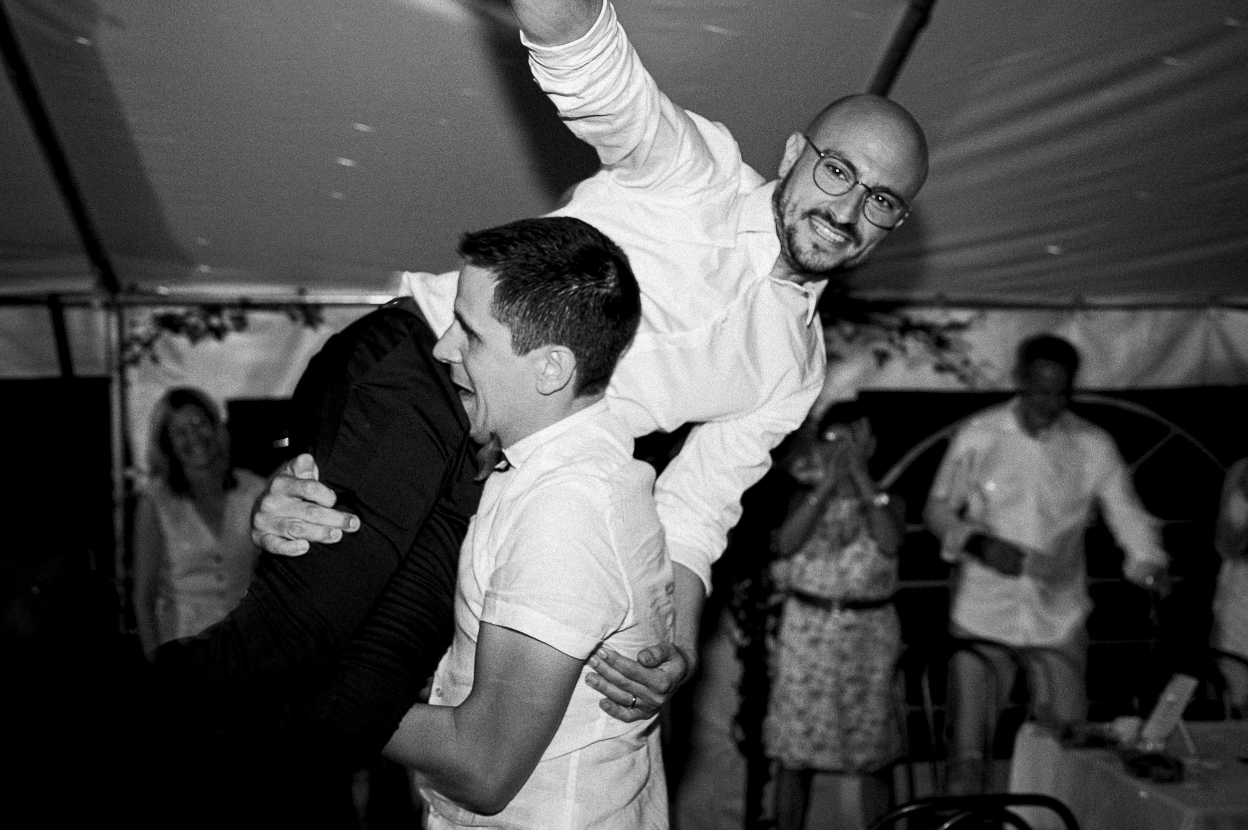 Deux hommes souriants dansent lors d'une fête, en train de se tenir par la taille. L'un porte une chemise blanche, l'autre une veste noire. En arrière-plan, d'autres personnes participent à la fête dans un lieu décoré.