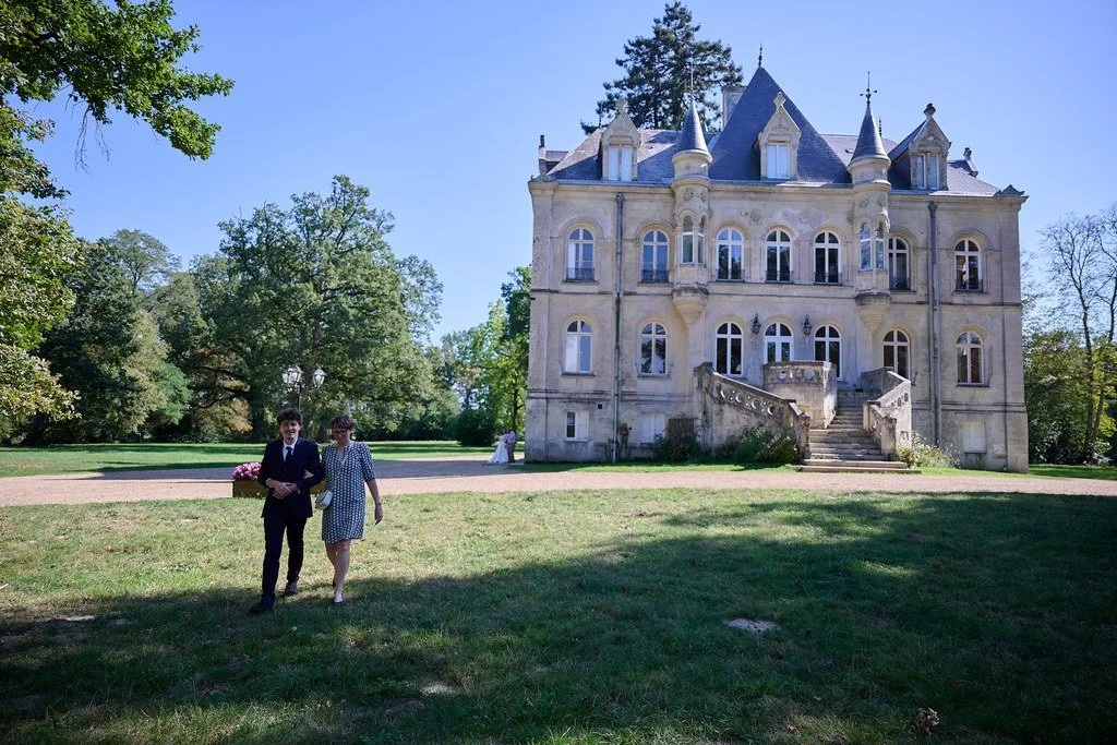 Deux personnes, un homme en costume et une femme en robe à pois, se promènent devant un grand château en pierre avec des tourelles, entouré d'arbres verts, par une journée ensoleillée.