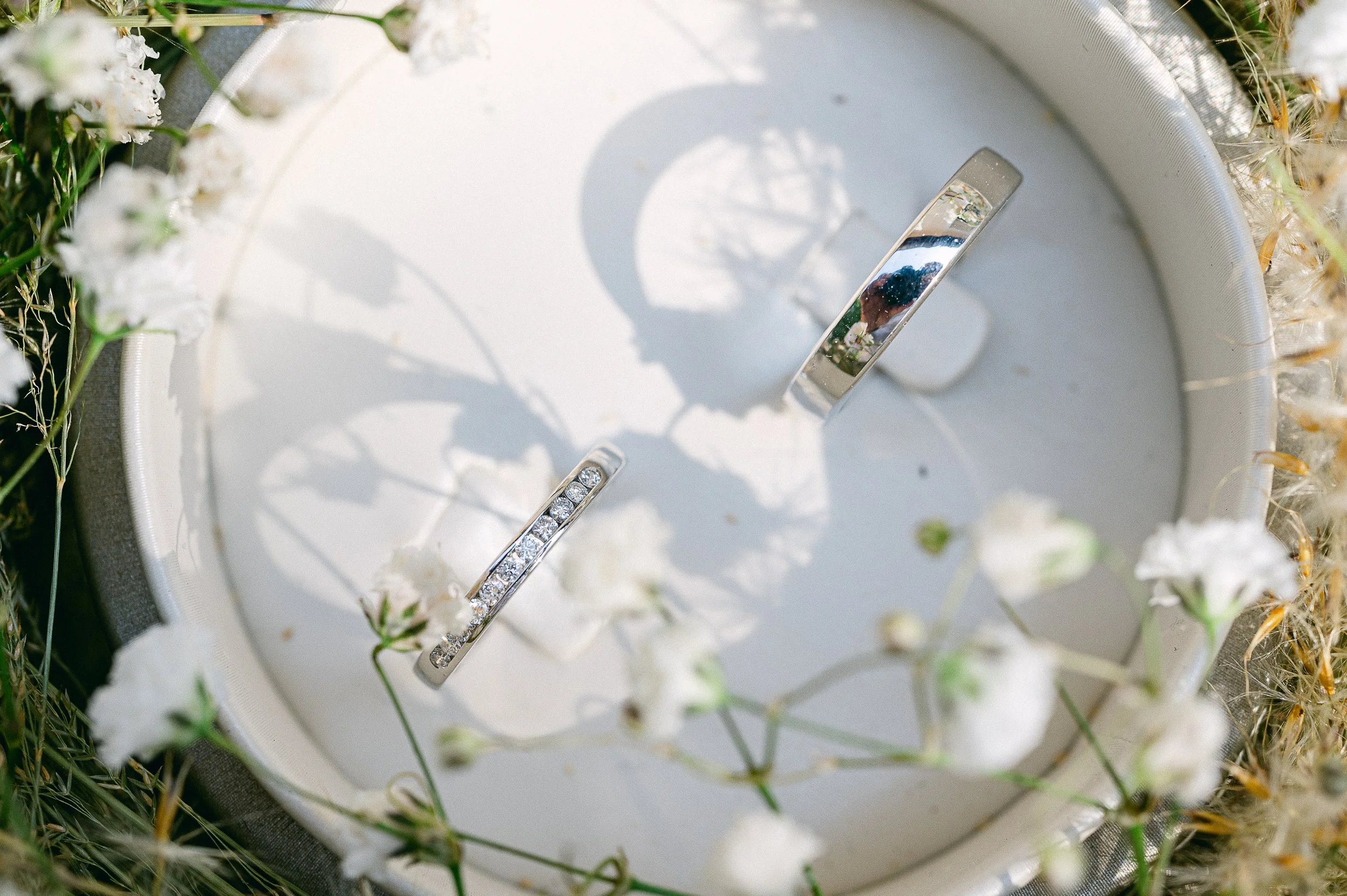 Deux bagues en argent avec des pierres précieuses reposant sur un lit de fleurs blanches, entourées de petites abeilles.