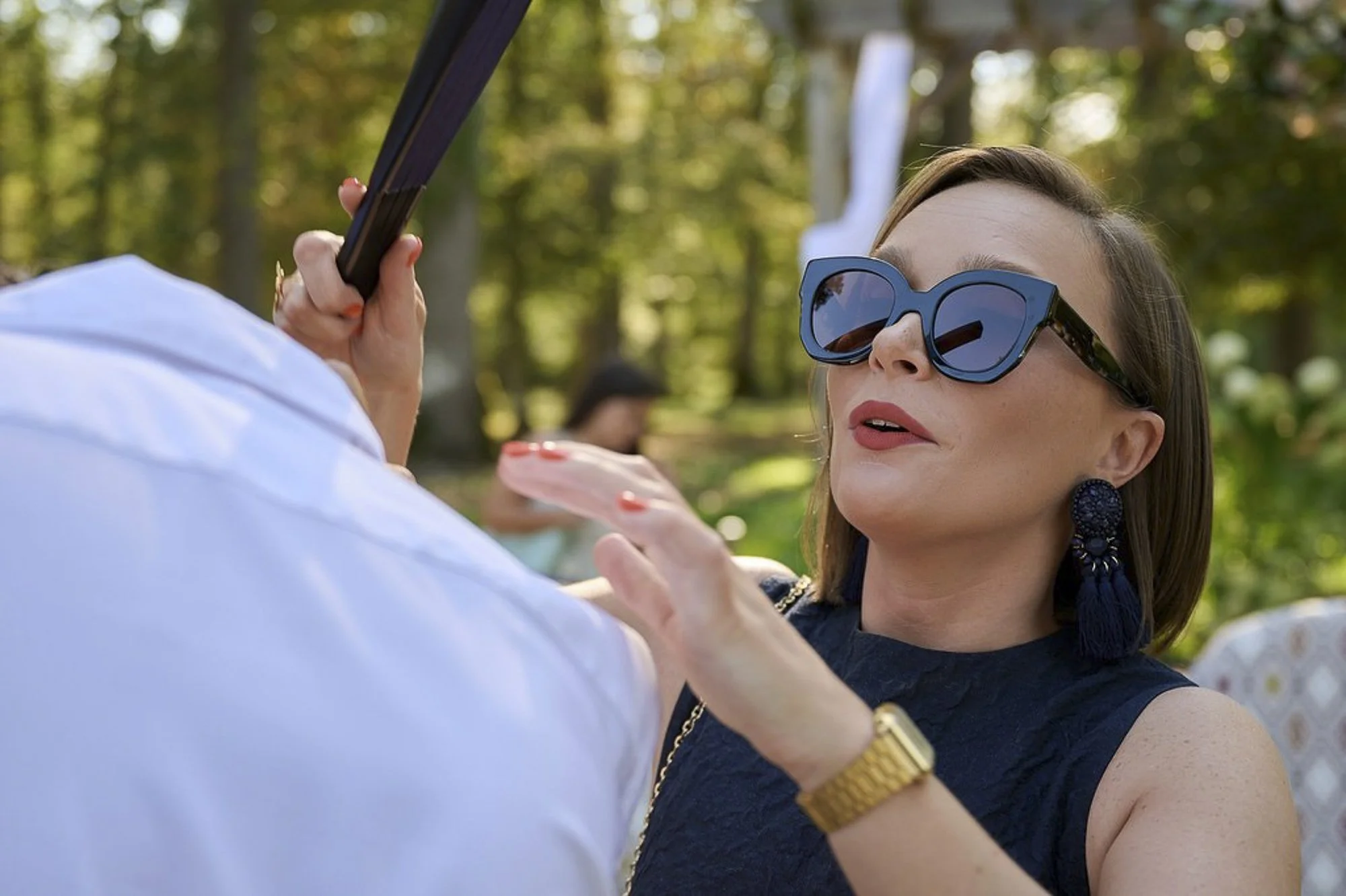 Une femme portant des lunettes de soleil et des boucles d'oreilles noires formatrices montre une bague à une personne en tenue blanche dans un parc. Arrière-plan avec arbres et autres personnes.