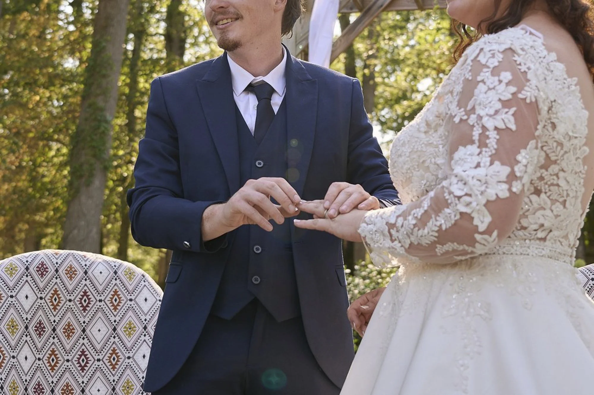 Un couple de mariés échangeant des vœux lors d'une cérémonie en plein air, avec un homme en costume bleu marine et une femme en robe de mariage avec broderies florales, entourés d'arbres en automne.