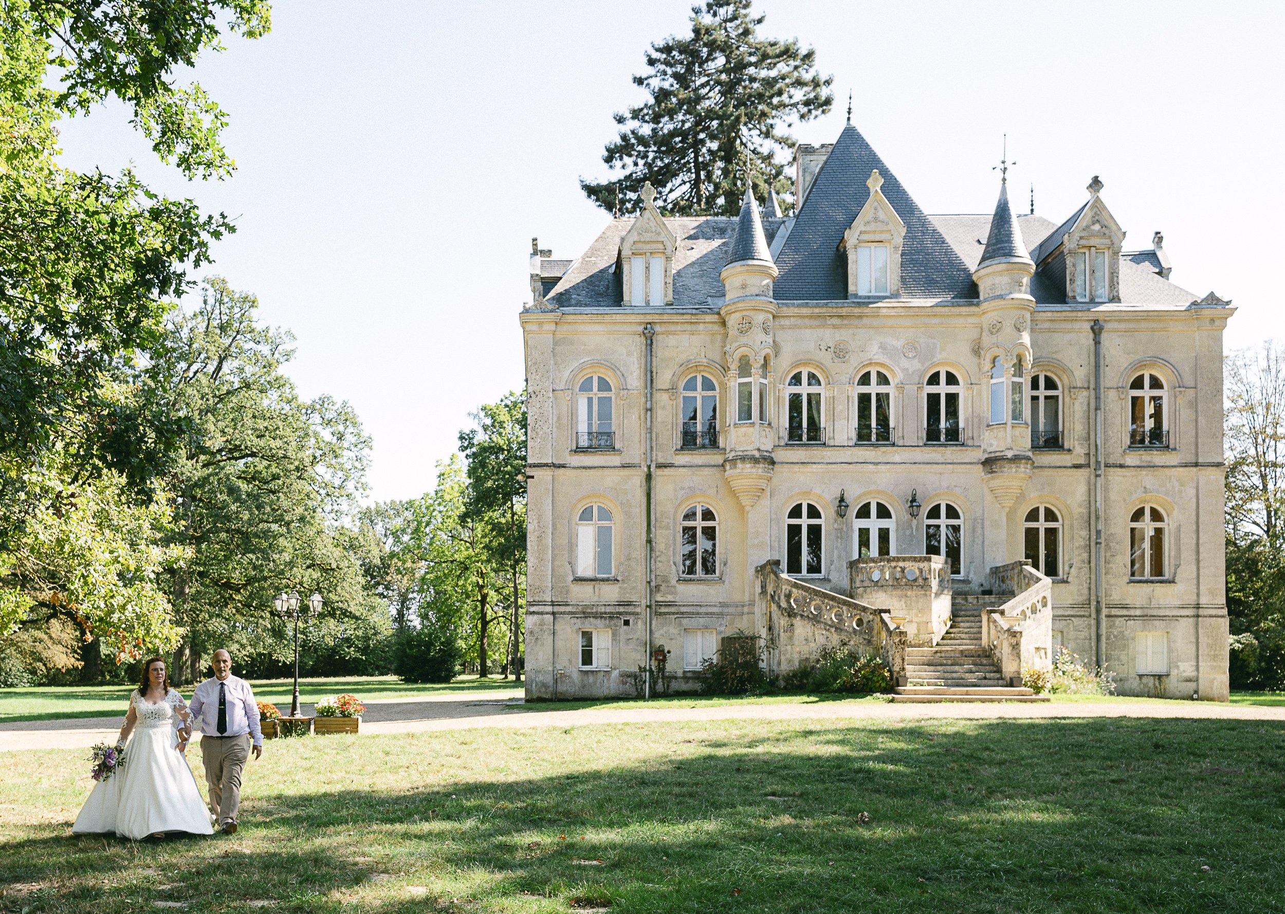 Un couple en mariage marche sur l'herbe devant un grand château en pierre, entouré d'arbres.