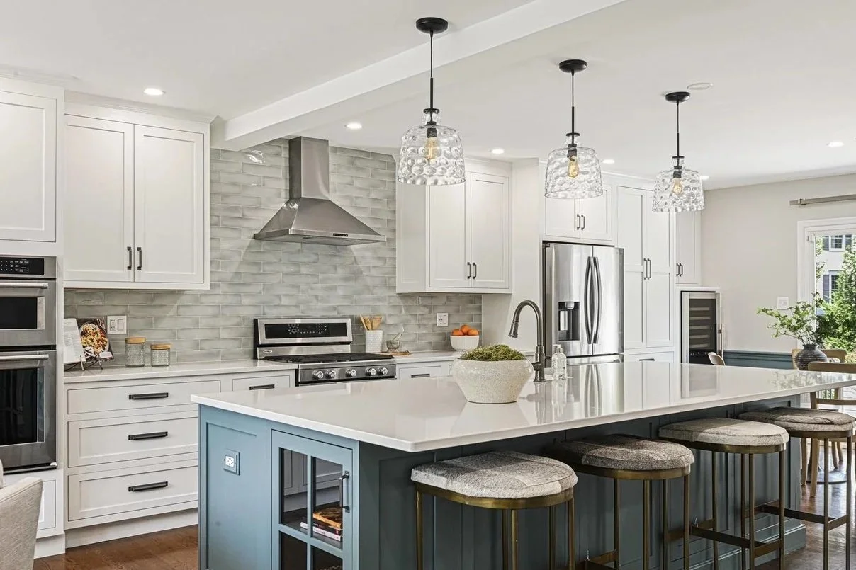 Modern kitchen with white cabinets, stainless steel appliances, a gray brick-style tile backsplash, and a blue island with a white countertop, decorated with pendant lights and bar stools.