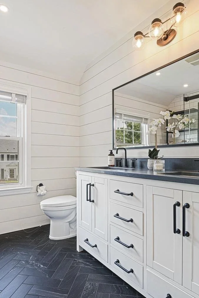 Modern bathroom with white shiplap walls, black hardware on a white vanity, and a large mirror. There is a window with blinds, a toilet, and a decorative white orchid in a glass vase. The ceiling has industrial-style lighting fixtures, and the floor is herringbone black tile.